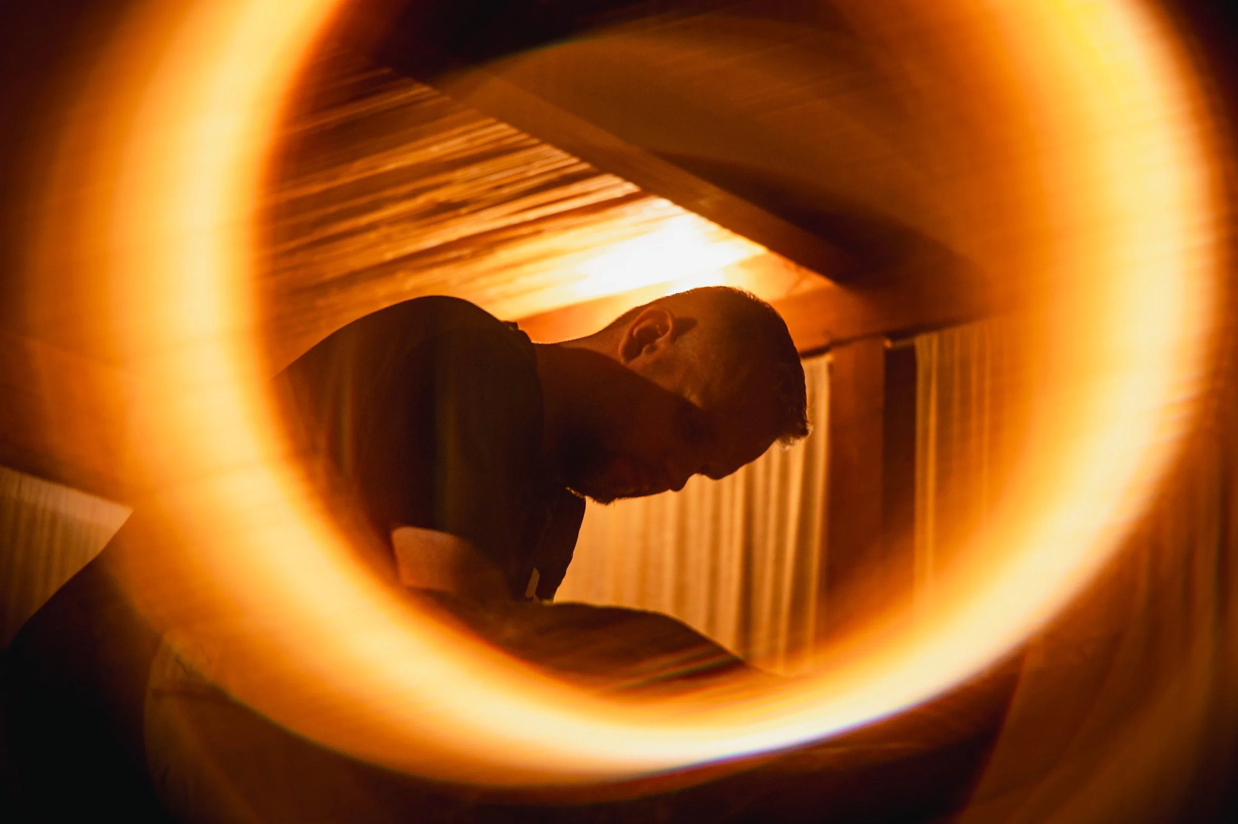 A person with short hair leaning over a surface in a dimly lit room with warm lighting, viewed through a circular opening that creates a glowing border around the image.