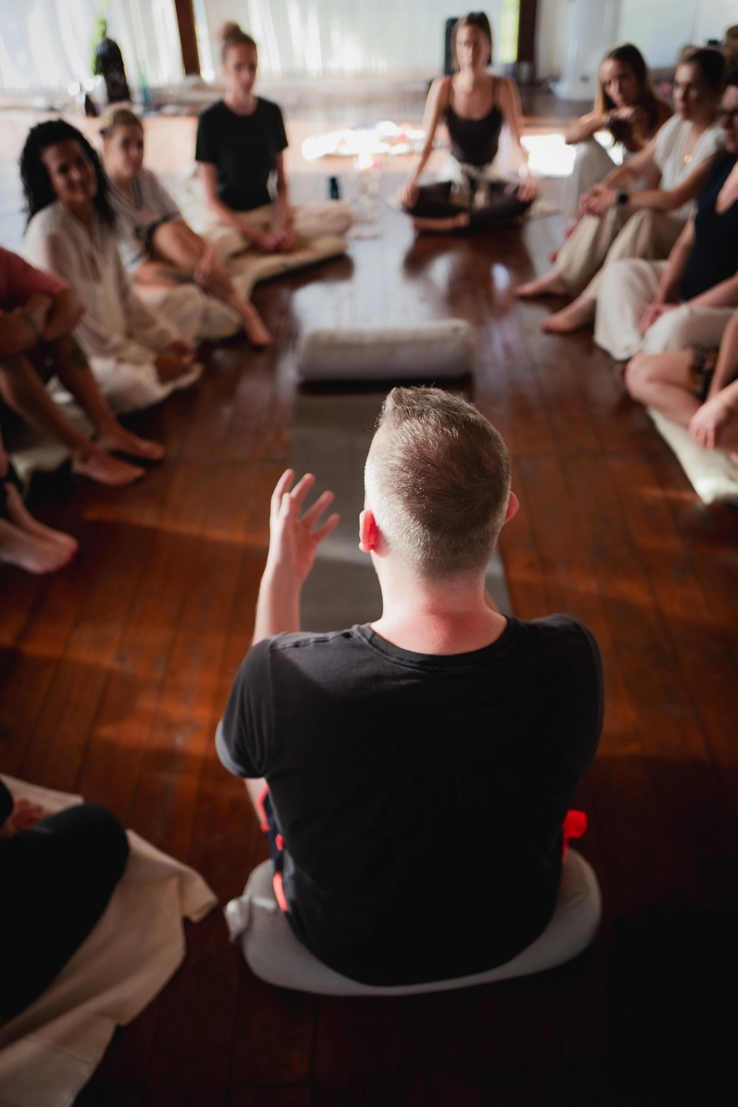 Person seated on the floor with their back to the camera, leading a group of people sitting in a circle on the floor.
