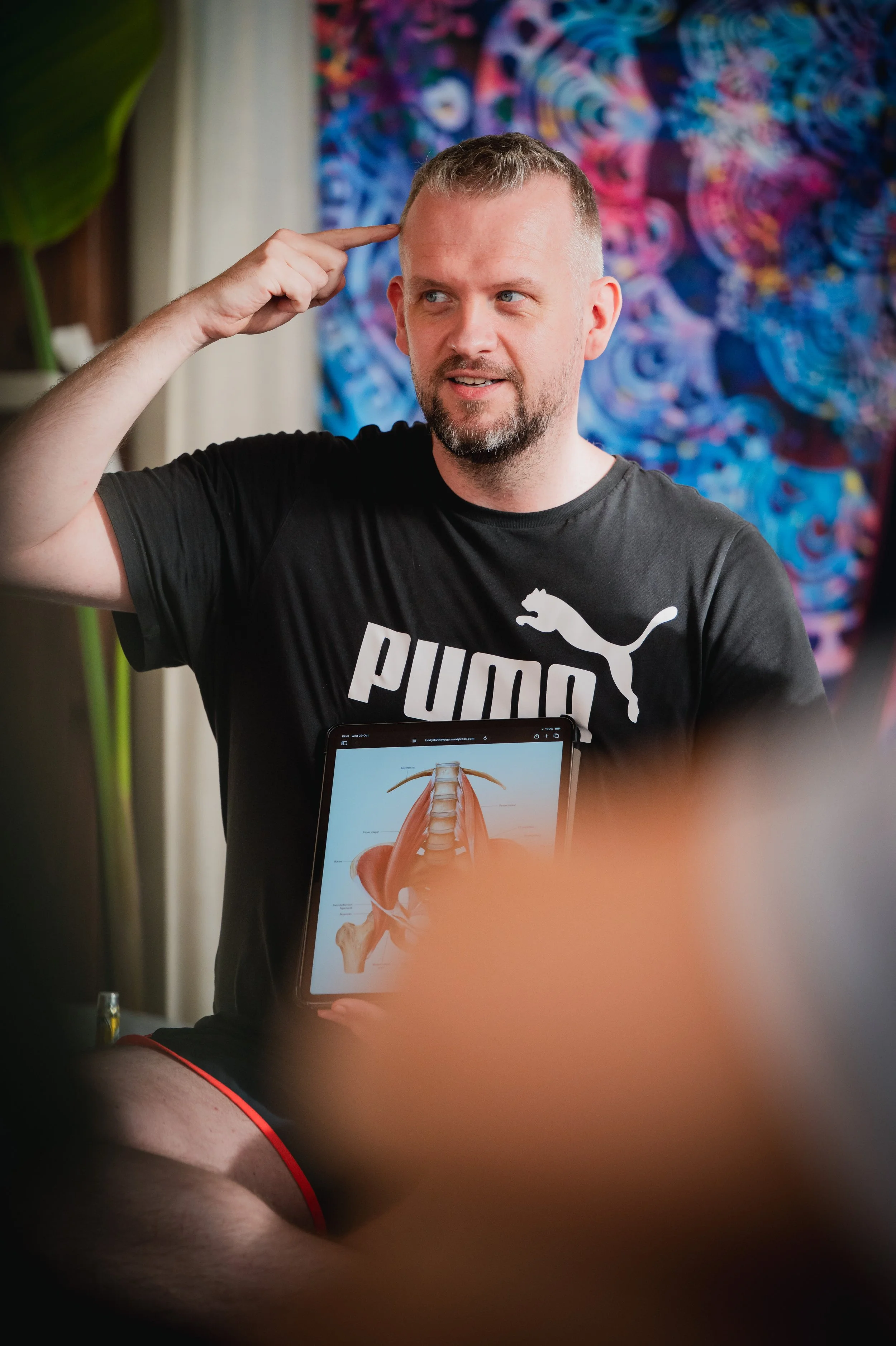 A man with short hair and a beard, wearing a black Puma T-shirt, gestures with his right hand to his temple while holding a tablet displaying a human anatomy diagram, in front of a colorful abstract background.