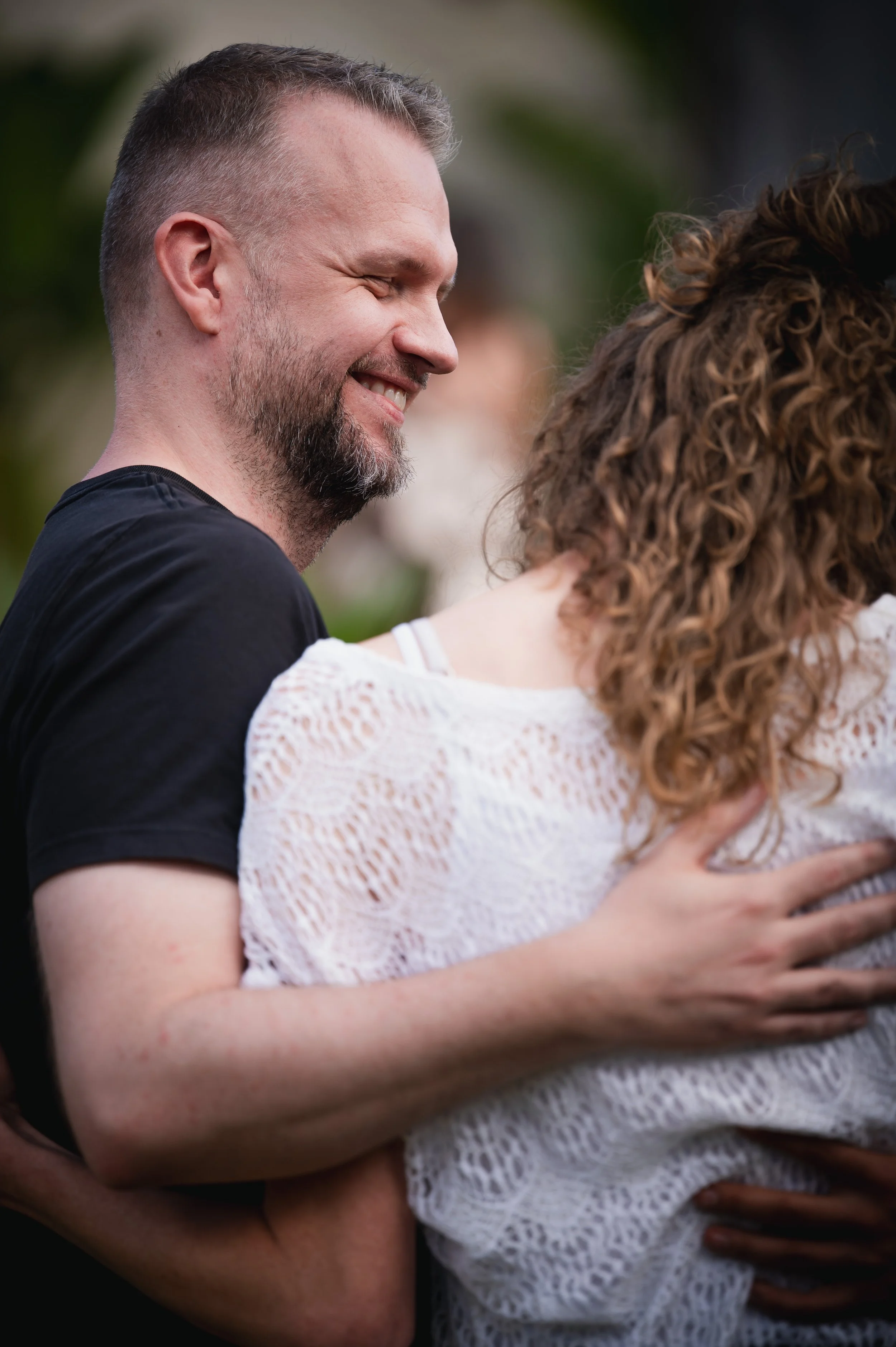 A man and a woman hugging, with the man smiling and eyes closed, outdoors with greenery in the background.