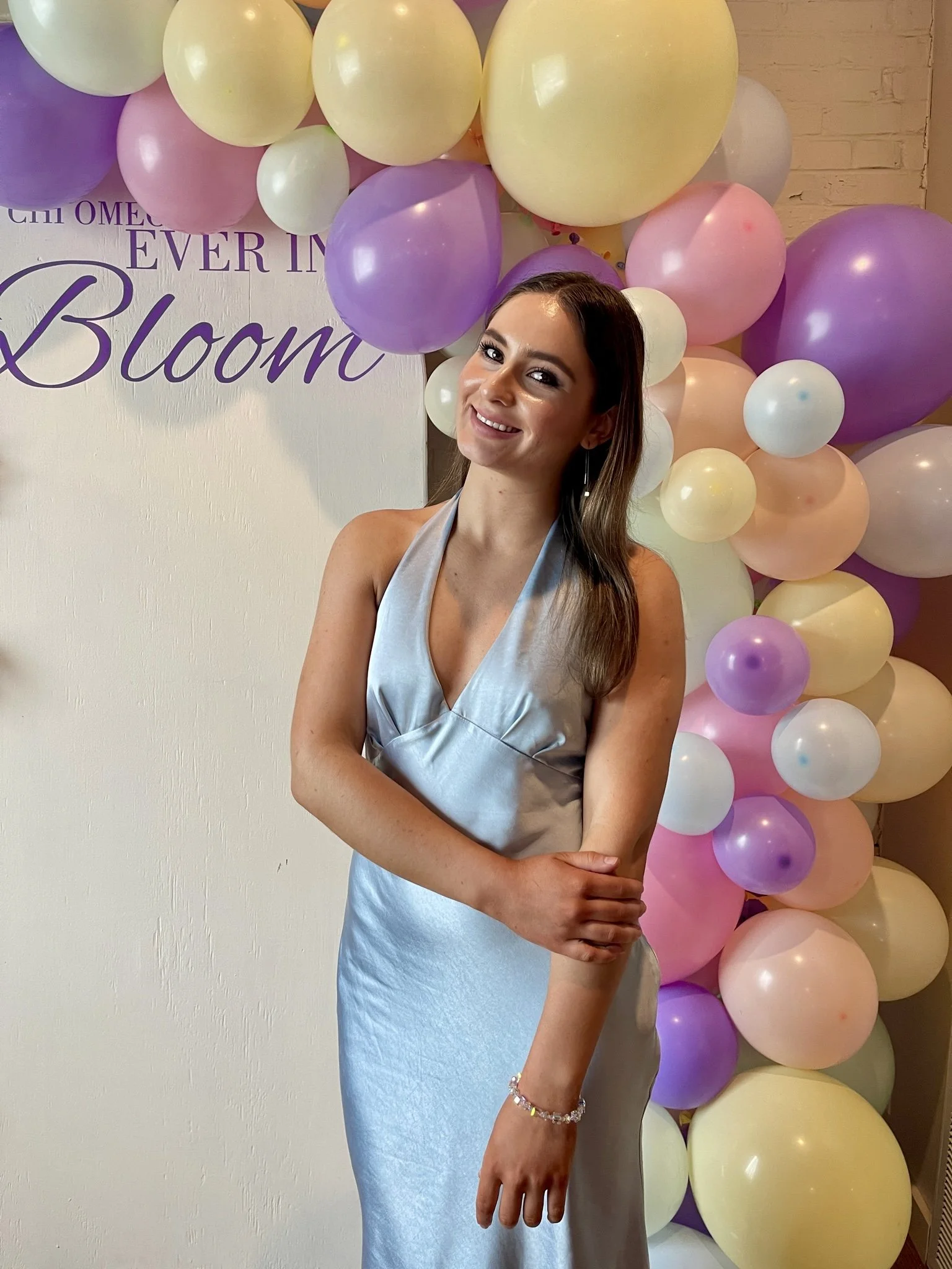 A woman standing in front of a backdrop decorated with pastel-colored balloons, wearing a silky silver dress and smiling at the camera.