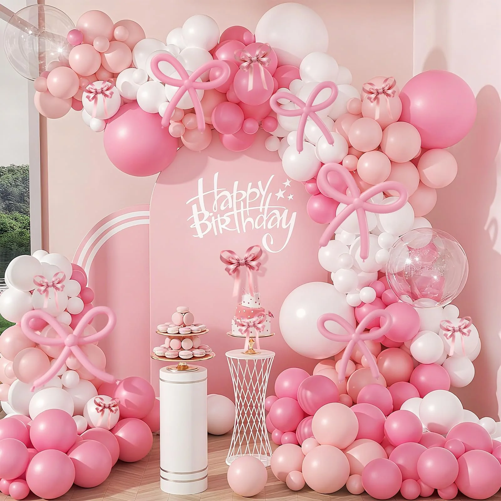 Decorative birthday party setup with pink, white, and clear balloons, pink bows, and a 'Happy Birthday' sign on a pink wall, with tables holding pink and white treats.