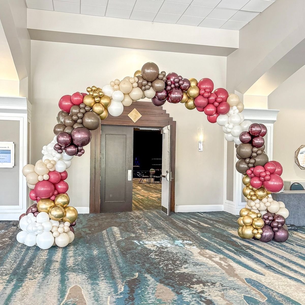 Grand opening balloon arch installation in Omaha featuring white, gold, shiny purple, brown, and pastel red balloons for storefront celebration.