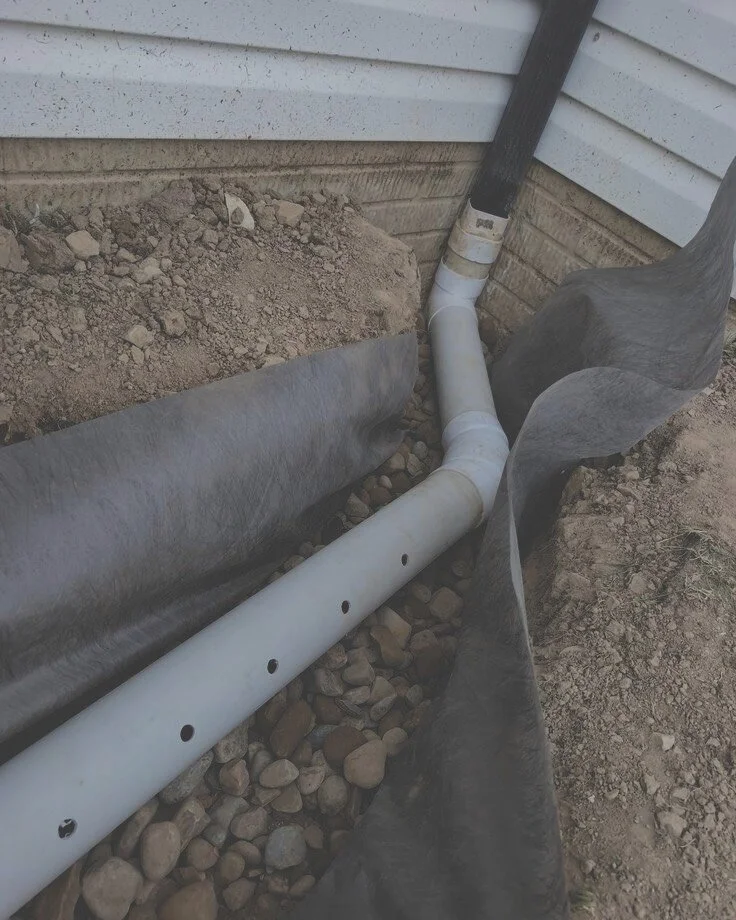 Underground drainage pipe installation next to the house foundation, with a protective black fabric liner and gravel surrounding the pipe.