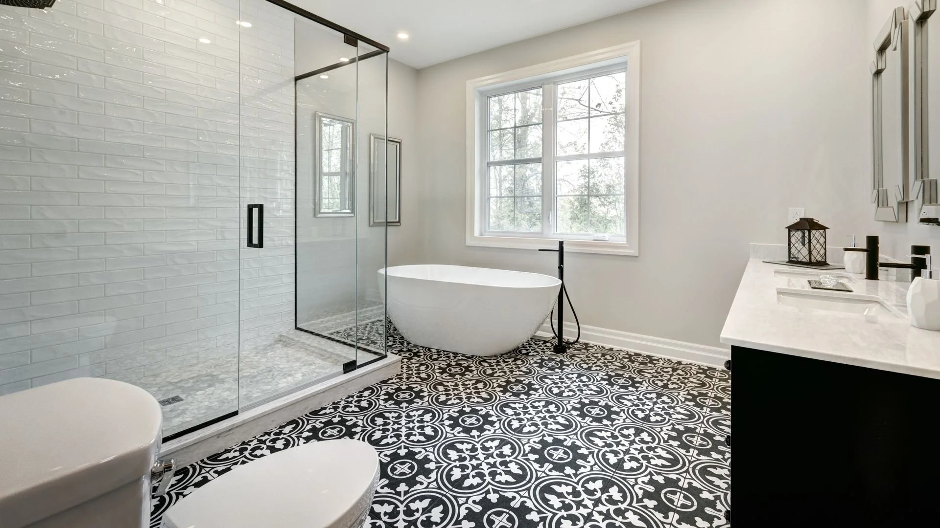 Modern bathroom featuring a walk-in glass shower with white subway tile, a free-standing bathtub near the window, a black faucet, and a double vanity with a white marble countertop and black fixtures, all on a decorative black and white patterned tile floor.
