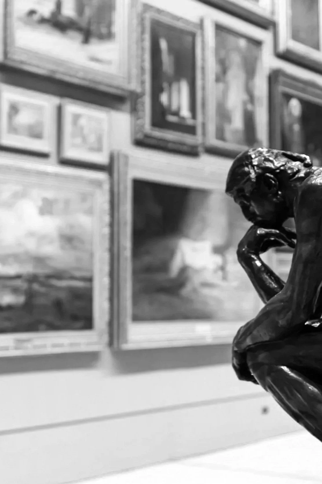 Meity Motif - a close-up of a black sculpture of a thinking man with a bust, in front of a wall with framed pictures or paintings. The sculpture's head is downward and hand resting on the chin.