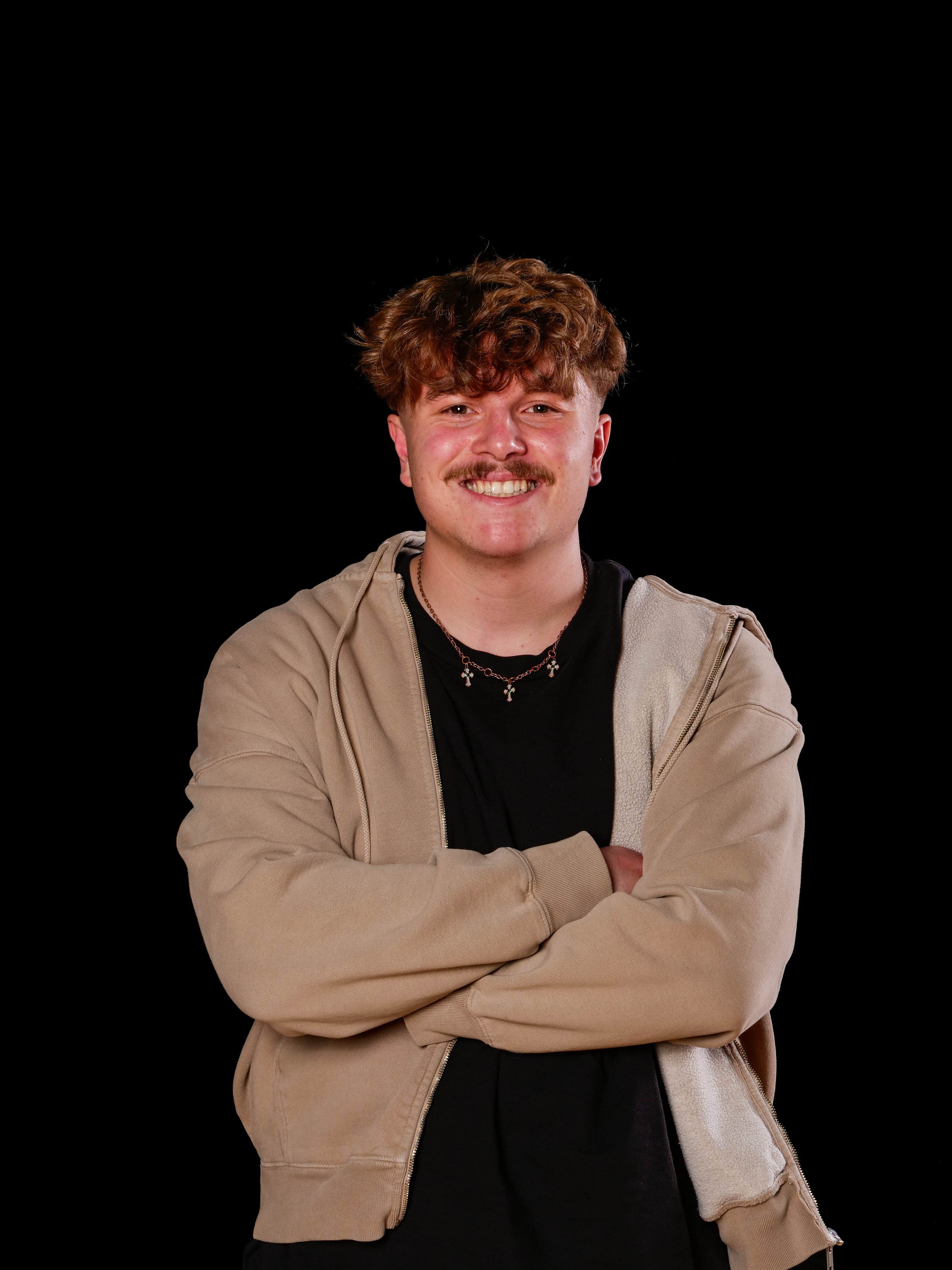 Portrait of Ethan Magnaghi, a graphic designer with curly brown hair and a mustache, smiling with arms crossed against a black background.
