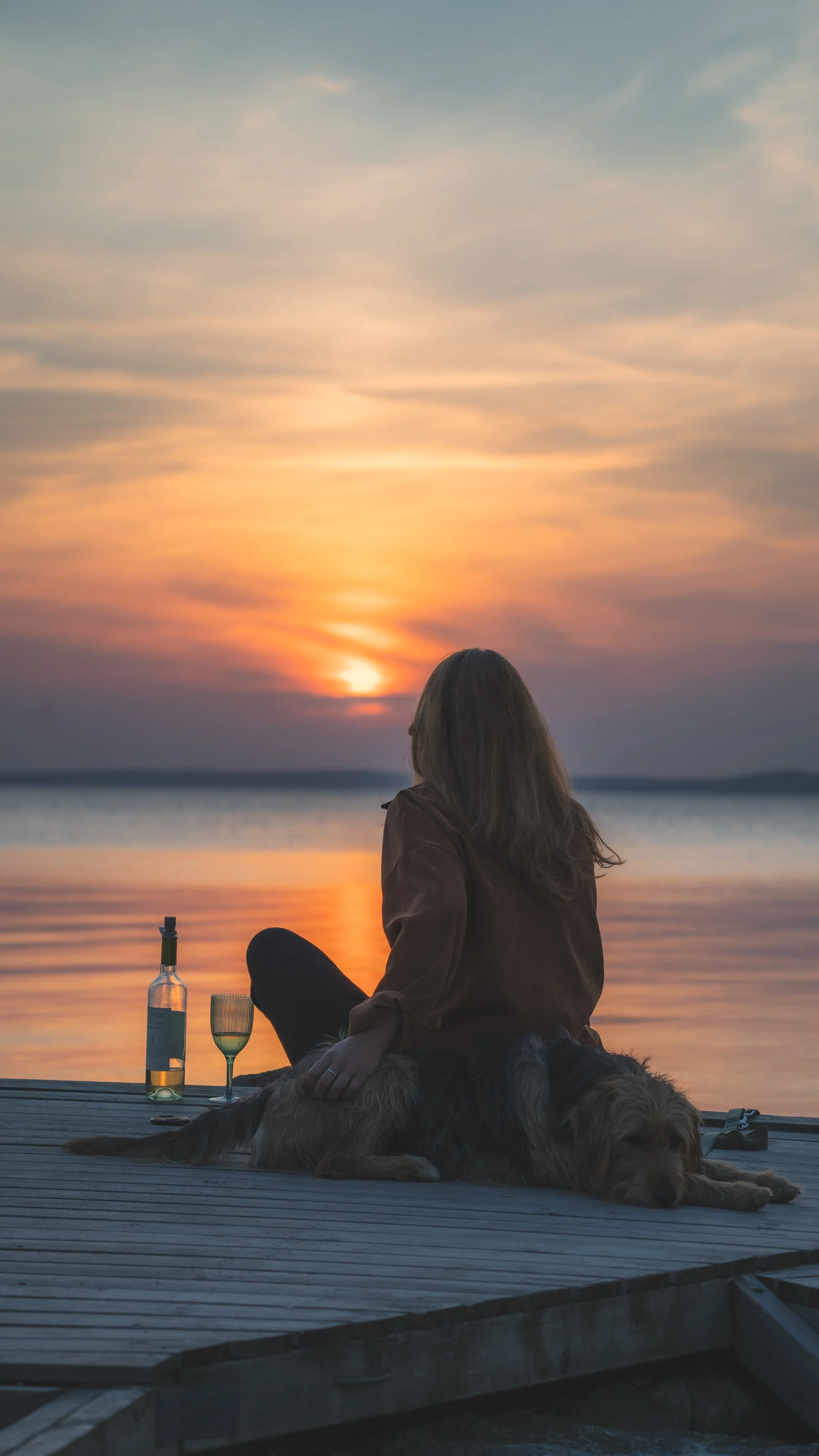 Eine Person sitzt auf einem Steg am Wasser bei Sonnenuntergang, begleitet von einem Hund. Es gibt eine Flasche Weisswein, ein Glas und eine Kerze vor der Person.