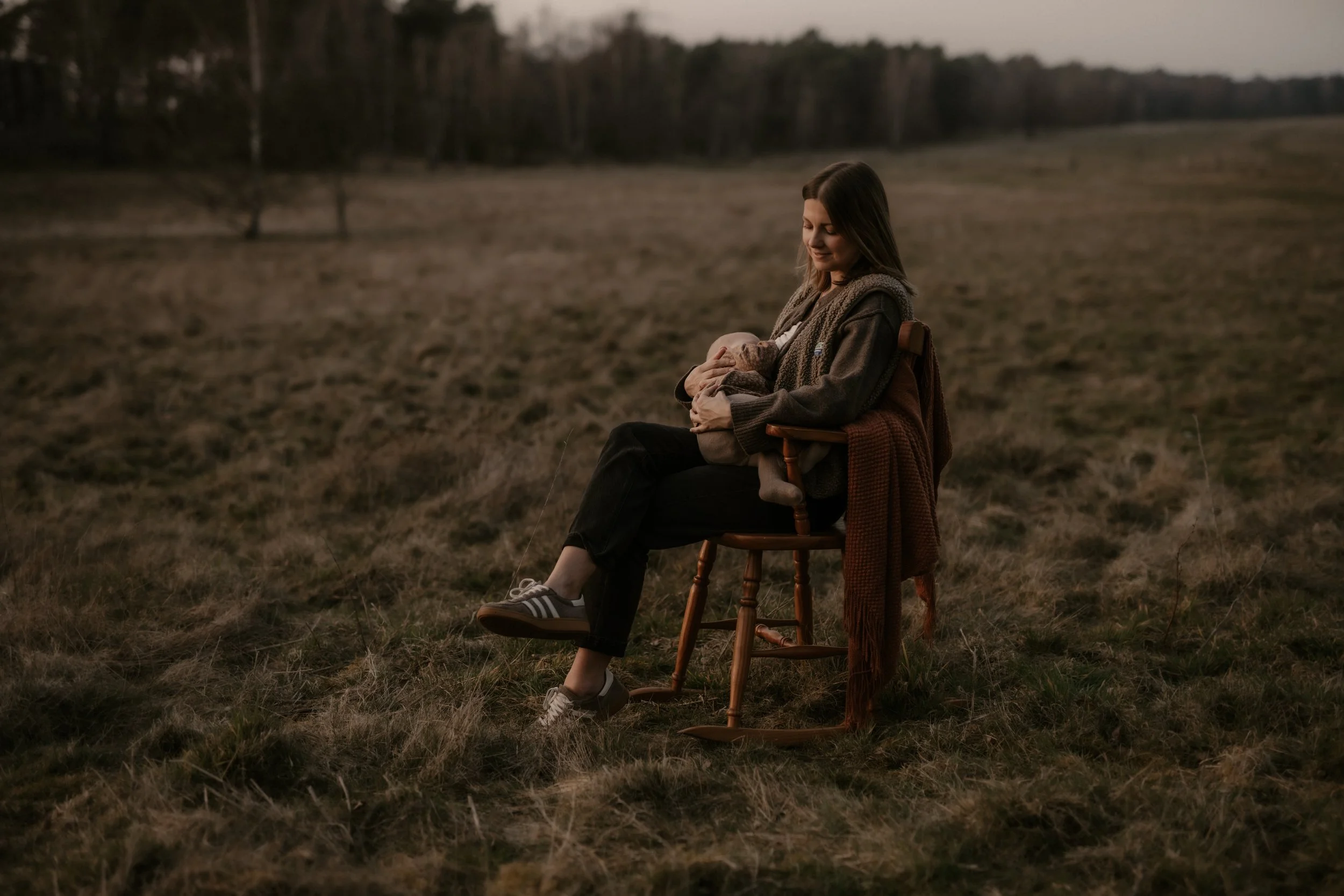 Eine Frau sitzt auf einem Holzstuhl im Freien auf einer Wiese, hält ein Baby und lächelt.