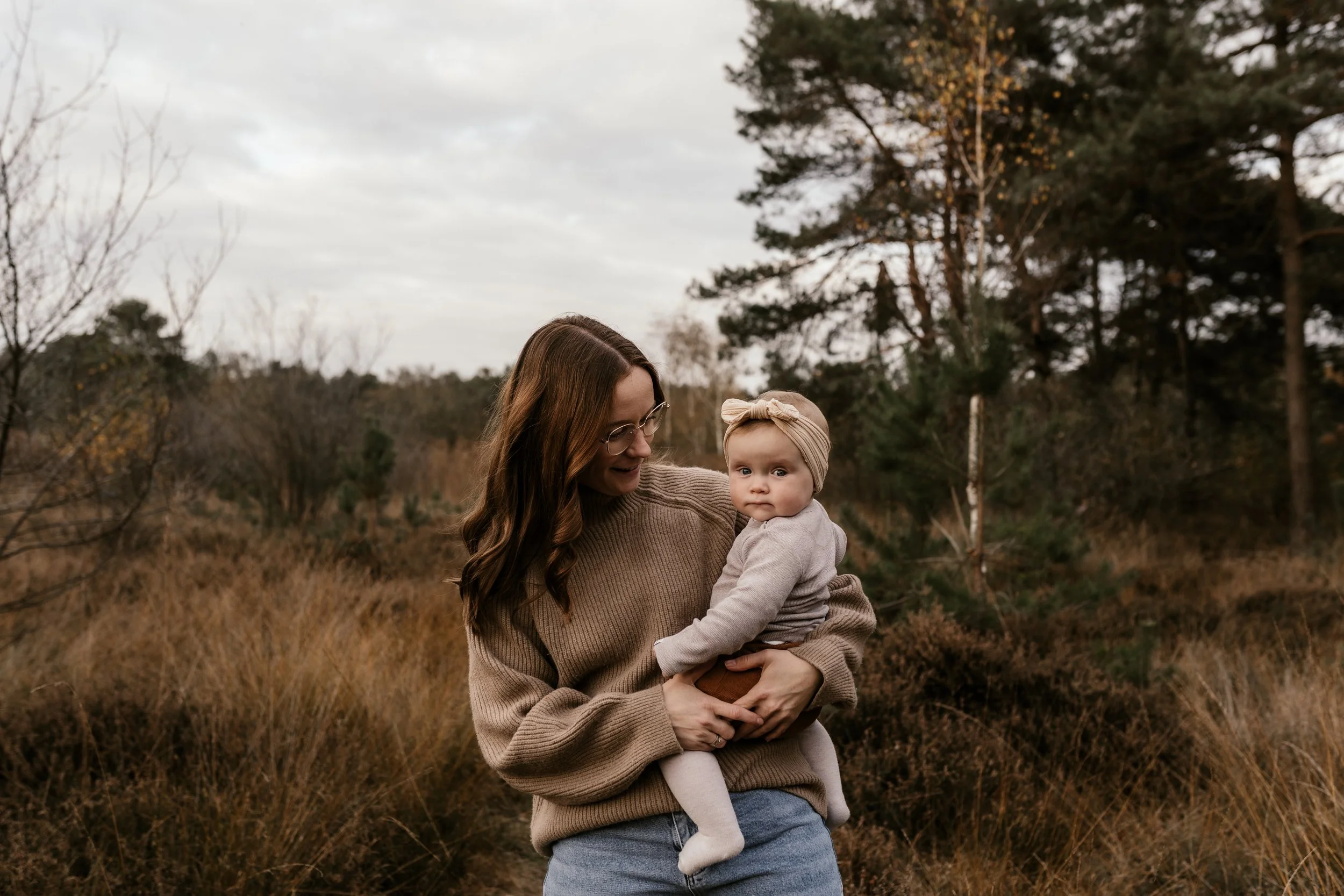 Eine Frau hält ein Baby im Freien auf einer Wiese mit Bäumen im Hintergrund, bewölkter Himmel.