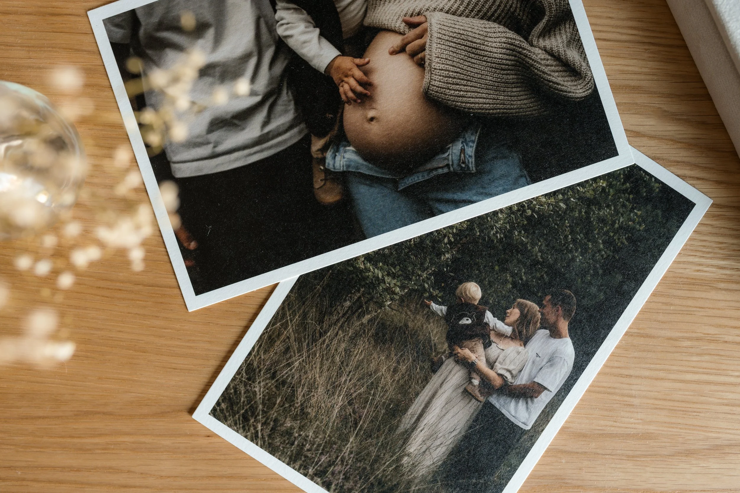 Zwei Fotografien auf einem Holztisch. Die obere zeigt eine schwangere Frau in Jeans und Strickpullover, die von jemand anderem berührt wird. Die untere zeigt eine Familie im Freien, ein Mann hält ein Kind im Arm, während eine Frau mit dem Kind in einer Naturkulisse steht.