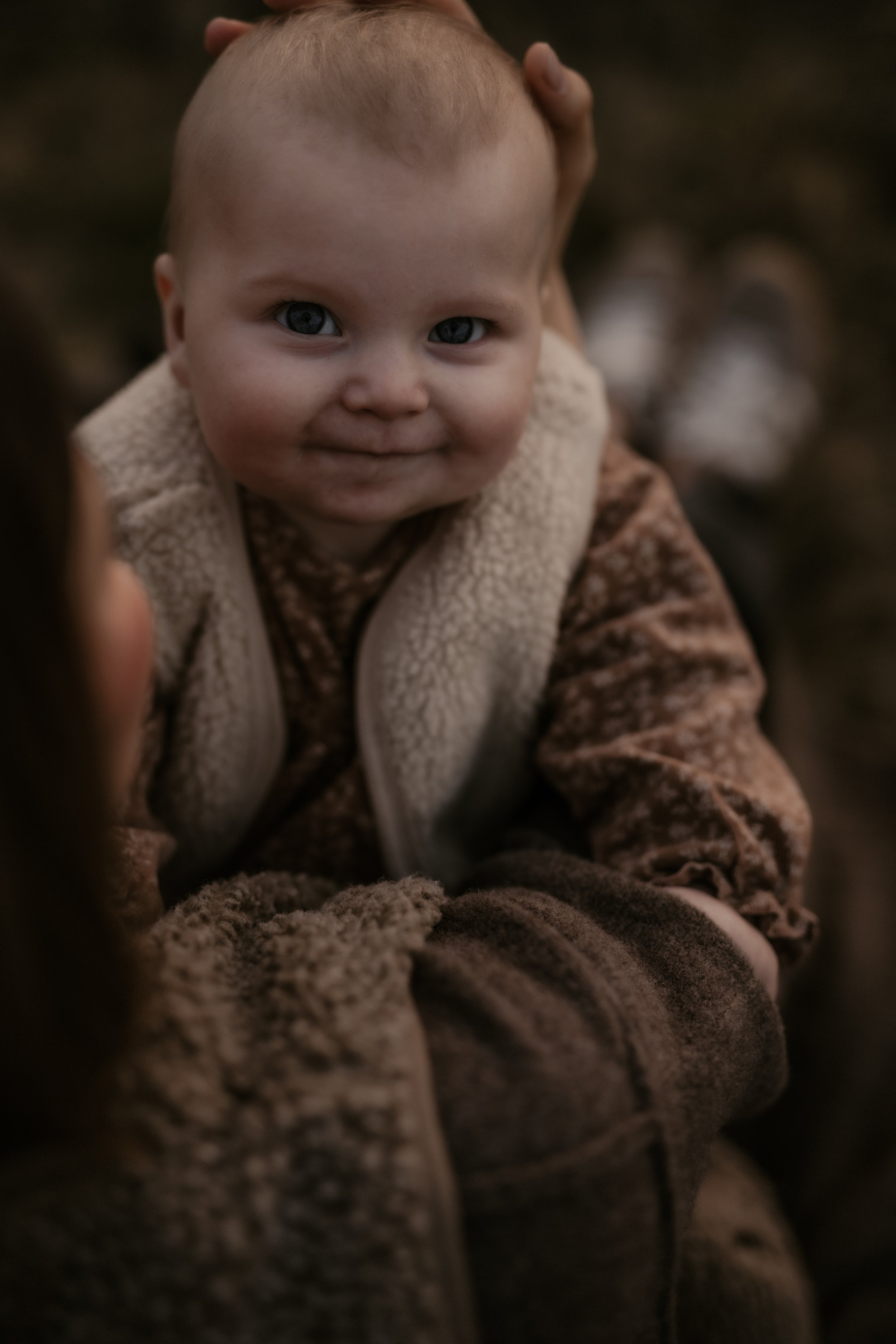 Ein Baby mit blauen Augen lächelt in die Kamera, gehalten von einer Person, die nur teilweise sichtbar ist.