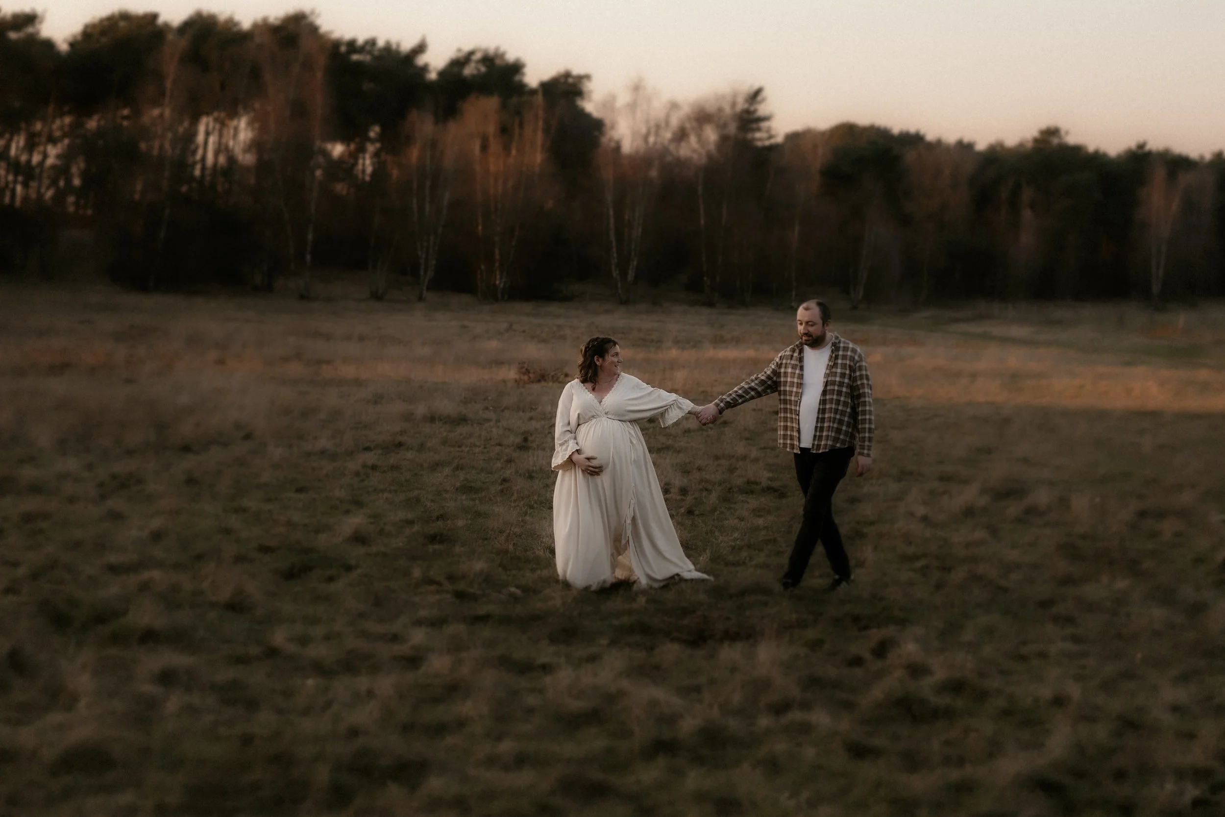 Ein Paar spaziert in einem offenen Feld bei Sonnenuntergang, der Mann hält die Hand der schwangeren Frau, die ein langes, helles Kleid trägt.