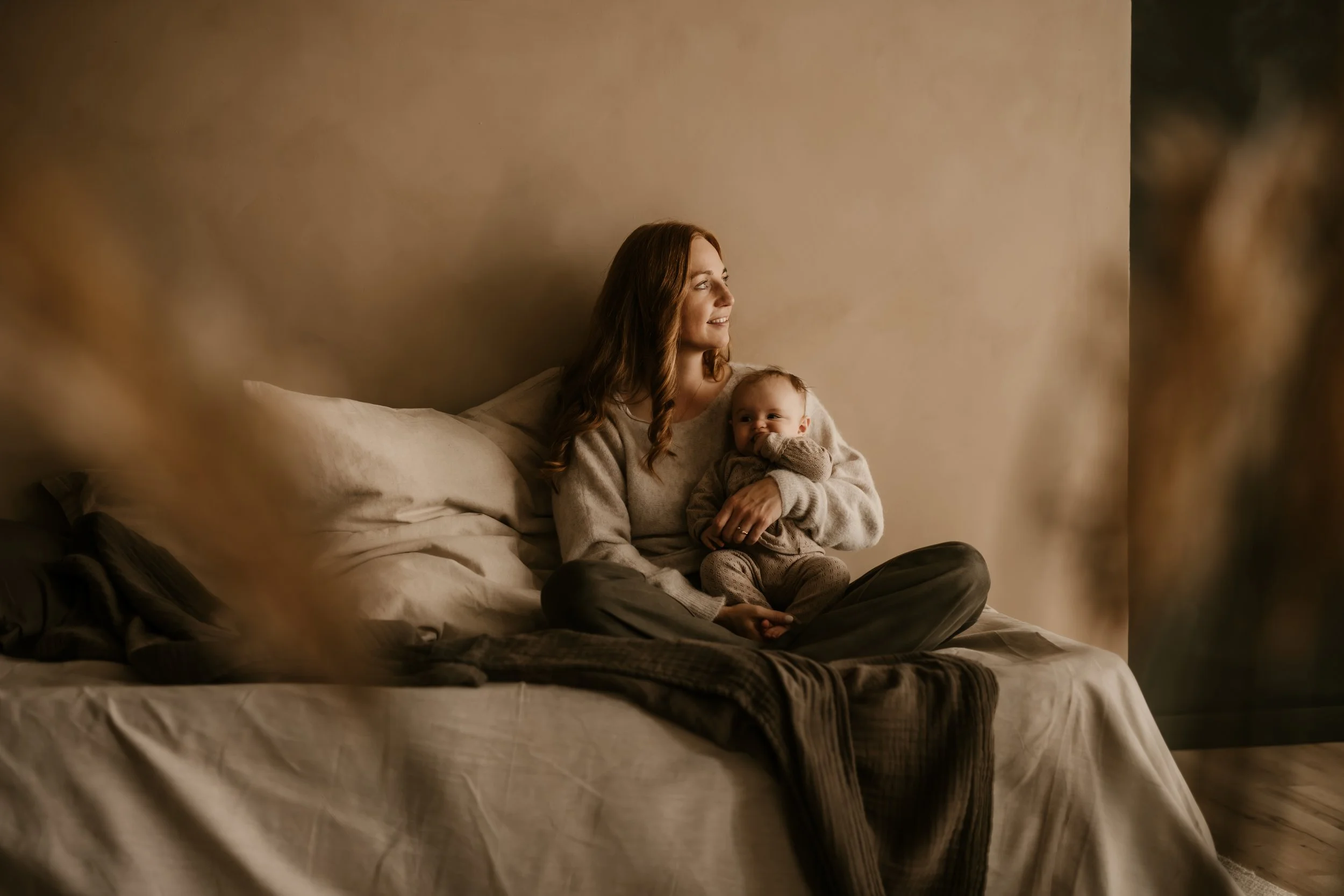 Eine Frau sitzt auf einem Bett und hält ein Baby im Arm, beide sehen entspannt aus, während sie nach draußen schauen.