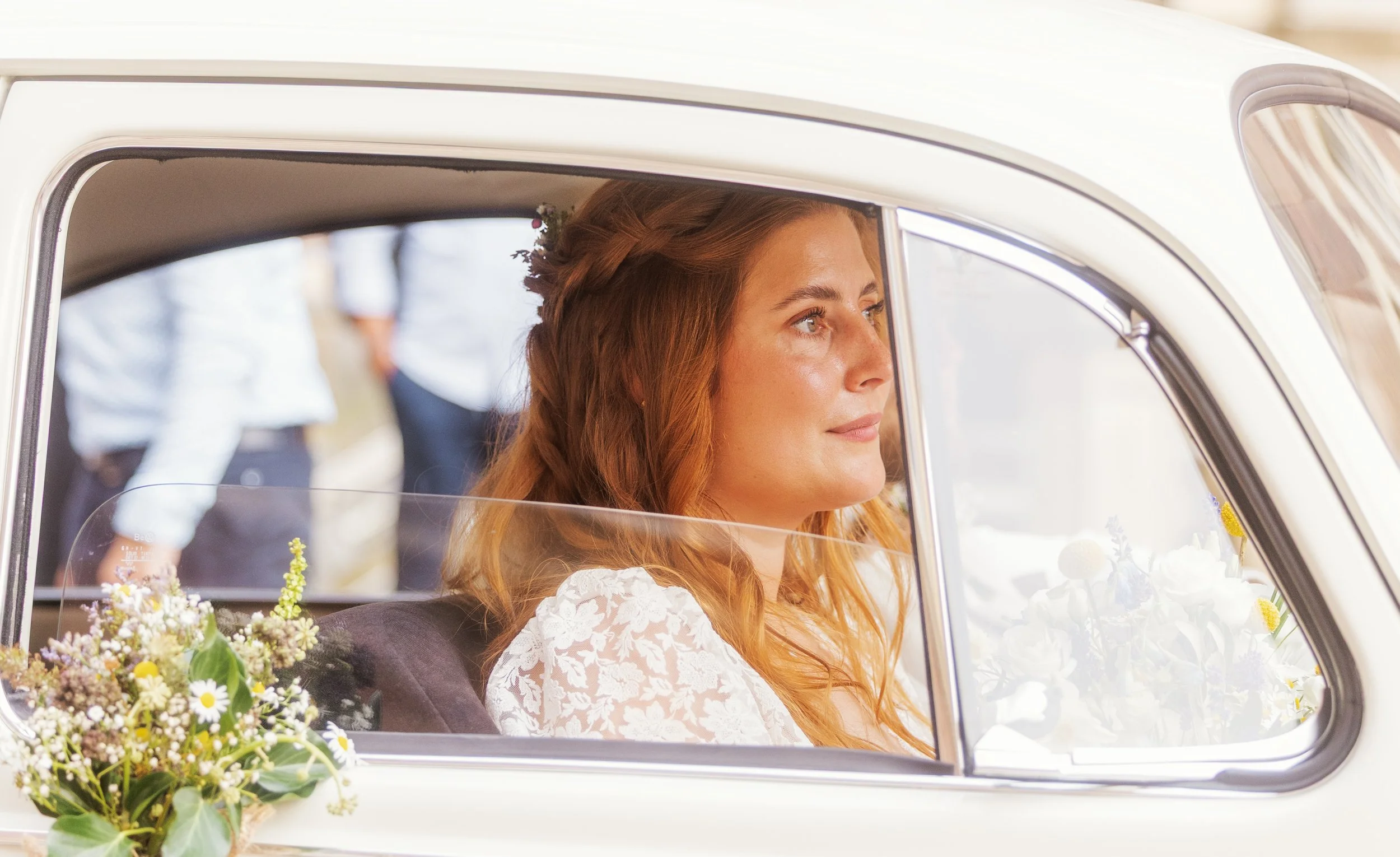 Braut mit roten Haaren sitzt in einem weißen Vintage-Auto, geschmückt mit Blumen, bei einer Hochzeit.