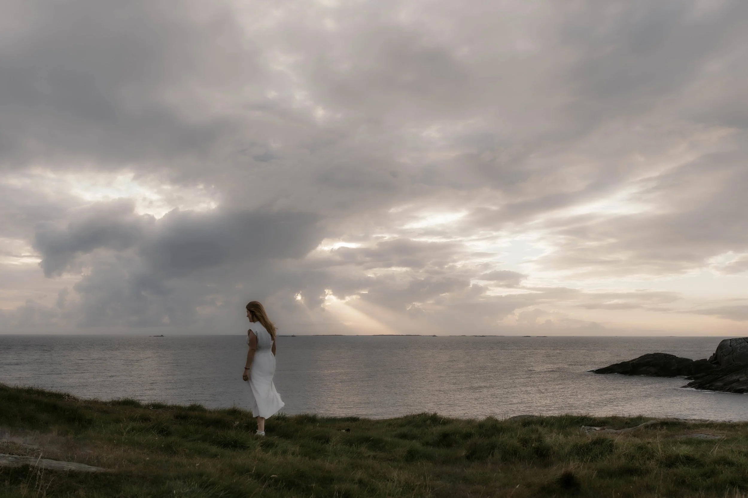Frau in weißem Kleid steht auf einer grünen Wiese am Meer bei bewölktem Himmel.