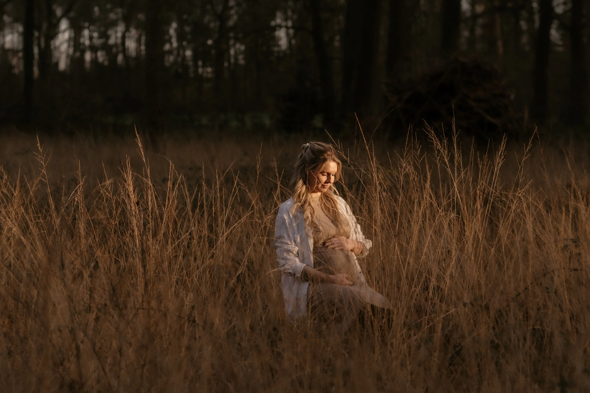 Eine schwangere Frau sitzt in einem trockenen Grasfeld im Abendlicht.