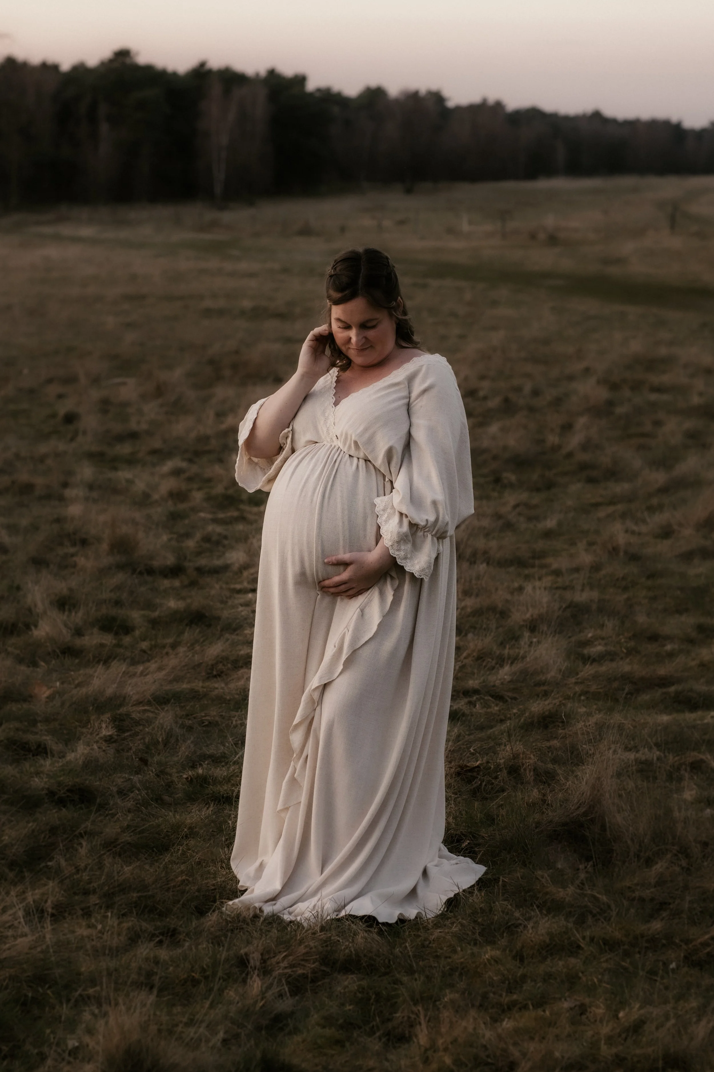 Eine schwangere Frau in einem weißen Kleid steht in einem offenen Feld bei Sonnenuntergang und hält ihre Hand auf ihren Bauch.