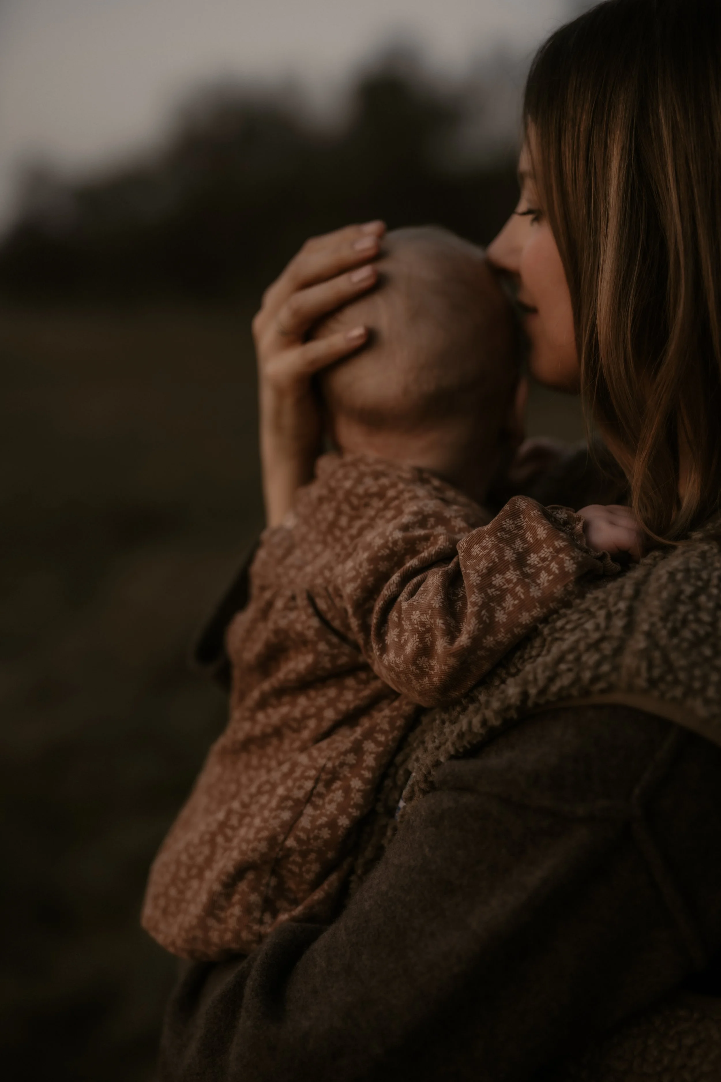 Eine Frau küsst ein Baby auf den Kopf, während sie es umarmt, im Freien bei Dämmerung.