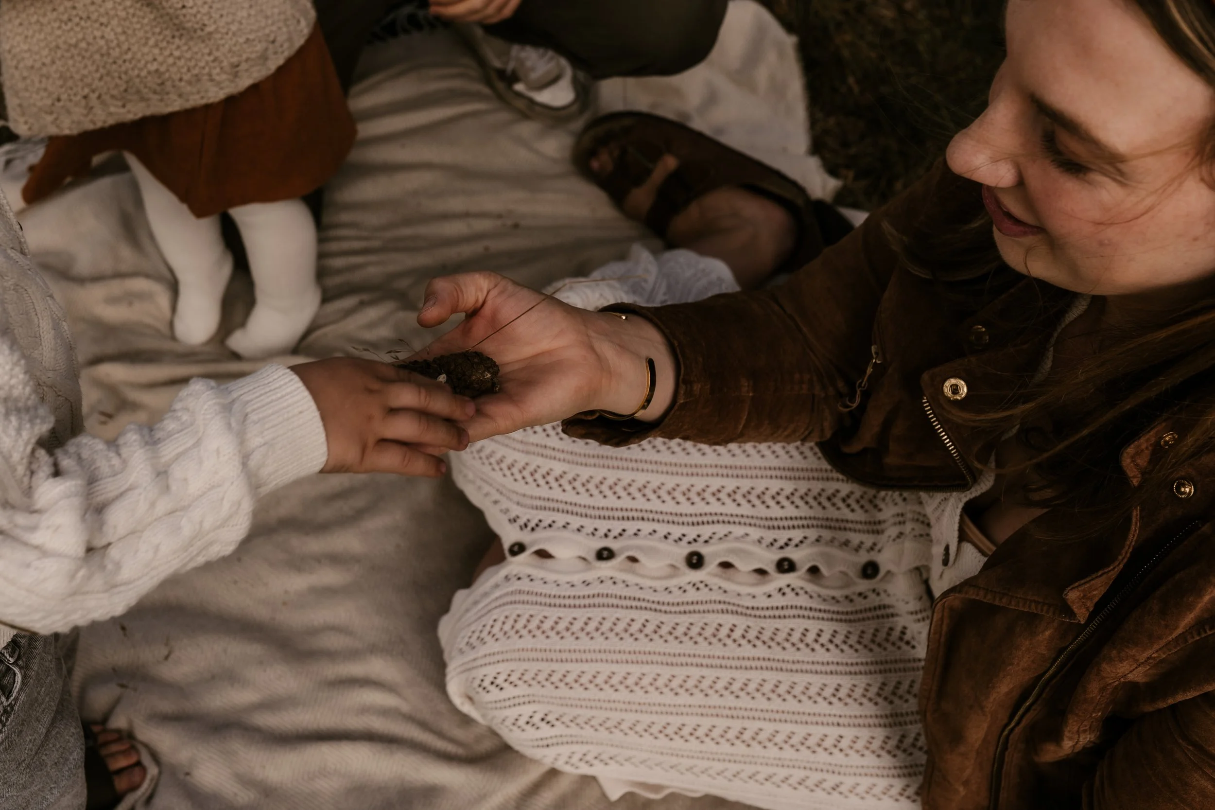 Eine junge Frau hält vorsichtig ein kleines, dunkles Tier in ihrer Hand, während ein Kind ebenfalls die Hand des Tieres berührt. Das Bild zeigt eine intime Szene, vermutlich in einem Innenraum, mit einem Baby im Hintergrund.