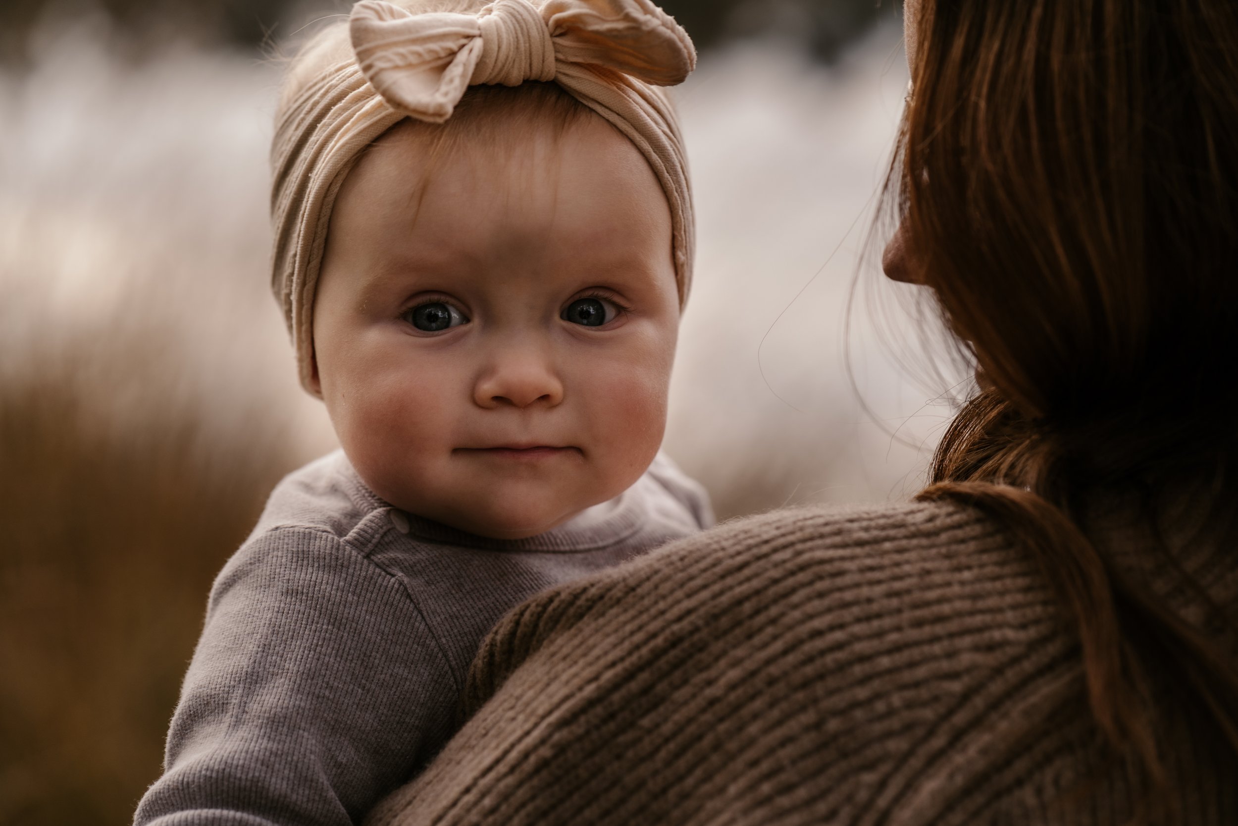 Ein Kleinkind mit einem rosafarbenen Stirnband, das auf einer Schulter einer Frau sitzt, in einer Naturumgebung mit Büschen und Bäumen im Hintergrund.