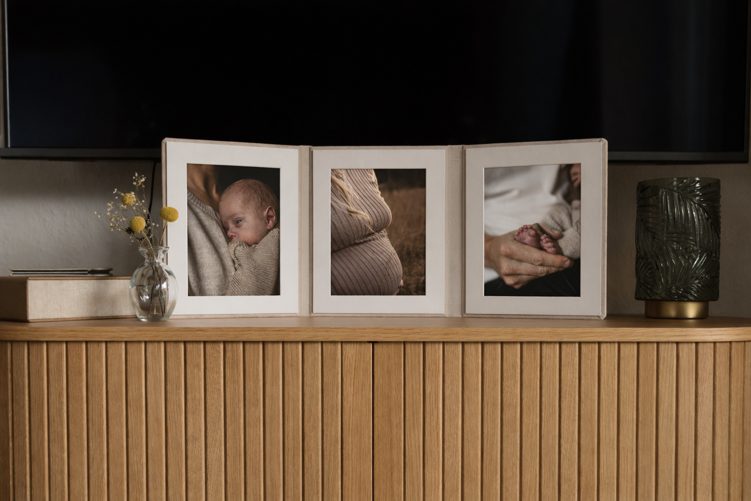 Drei Fotos in einem Rahmen auf einem Holzmöbel, eine zeigt ein Baby, das an die Brust einer Person gelehnt ist, die zweite einen Babybauch, und die dritte eine Hand, die ein Baby hält, alles in einem natürlichen Umfeld.