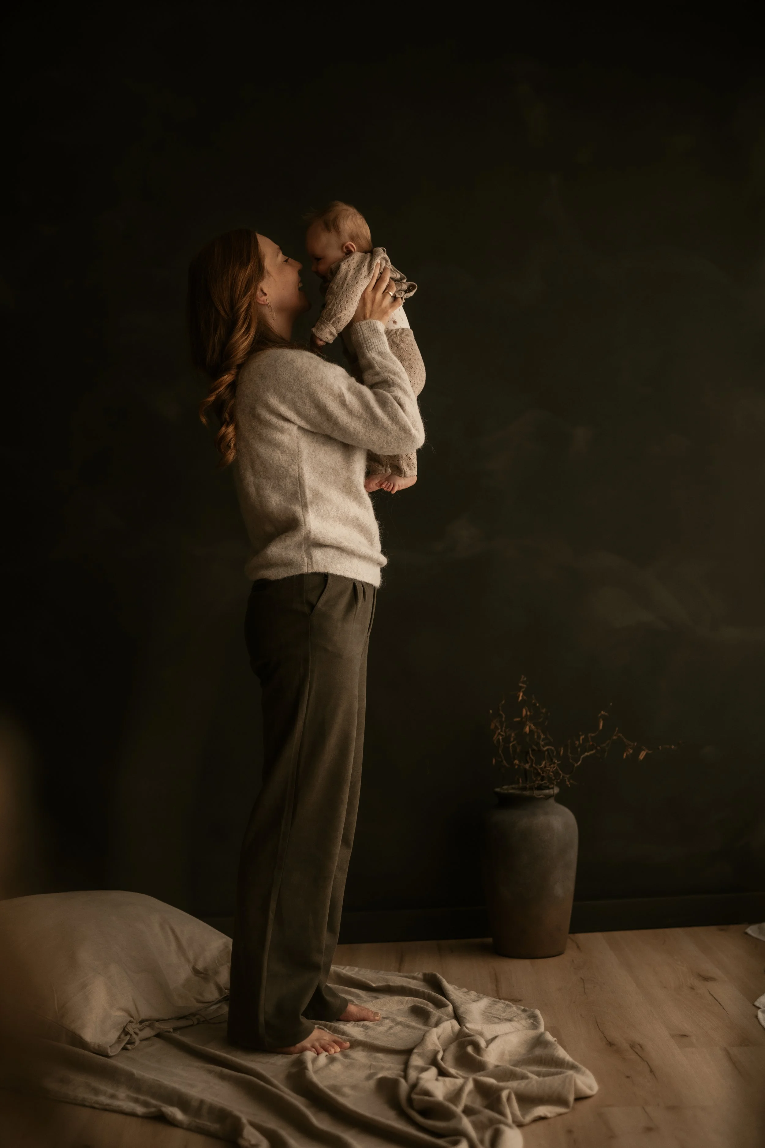Eine Frau hebt ein Baby hoch, beide lachen sich an, im Hintergrund eine dunkle Wand mit einer großen Vase, in der eine Pflanze oder Dekoration steht. Die Szenerie ist warm und ruhig, mit Kerzenschein.