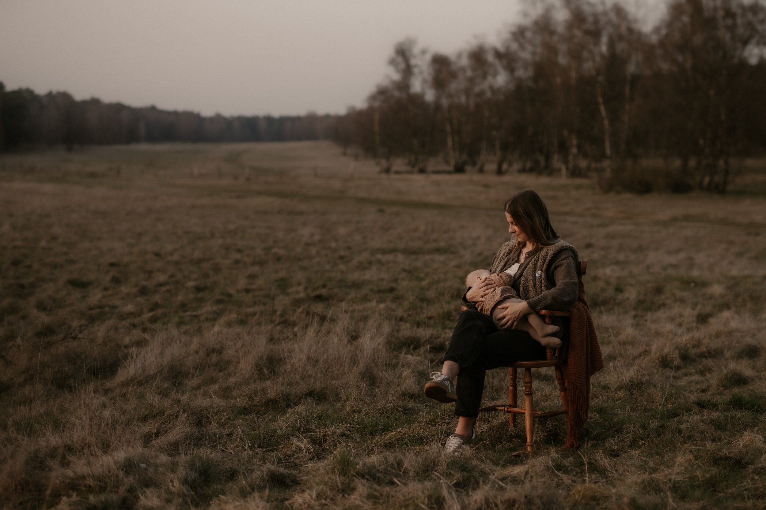 Eine Frau sitzt auf einem Holzstuhl in einer offenen Wiese und stillt ihr Baby, umgeben von einem weiten, leeren Feld mit Bäumen im Hintergrund bei Sonnenuntergang.