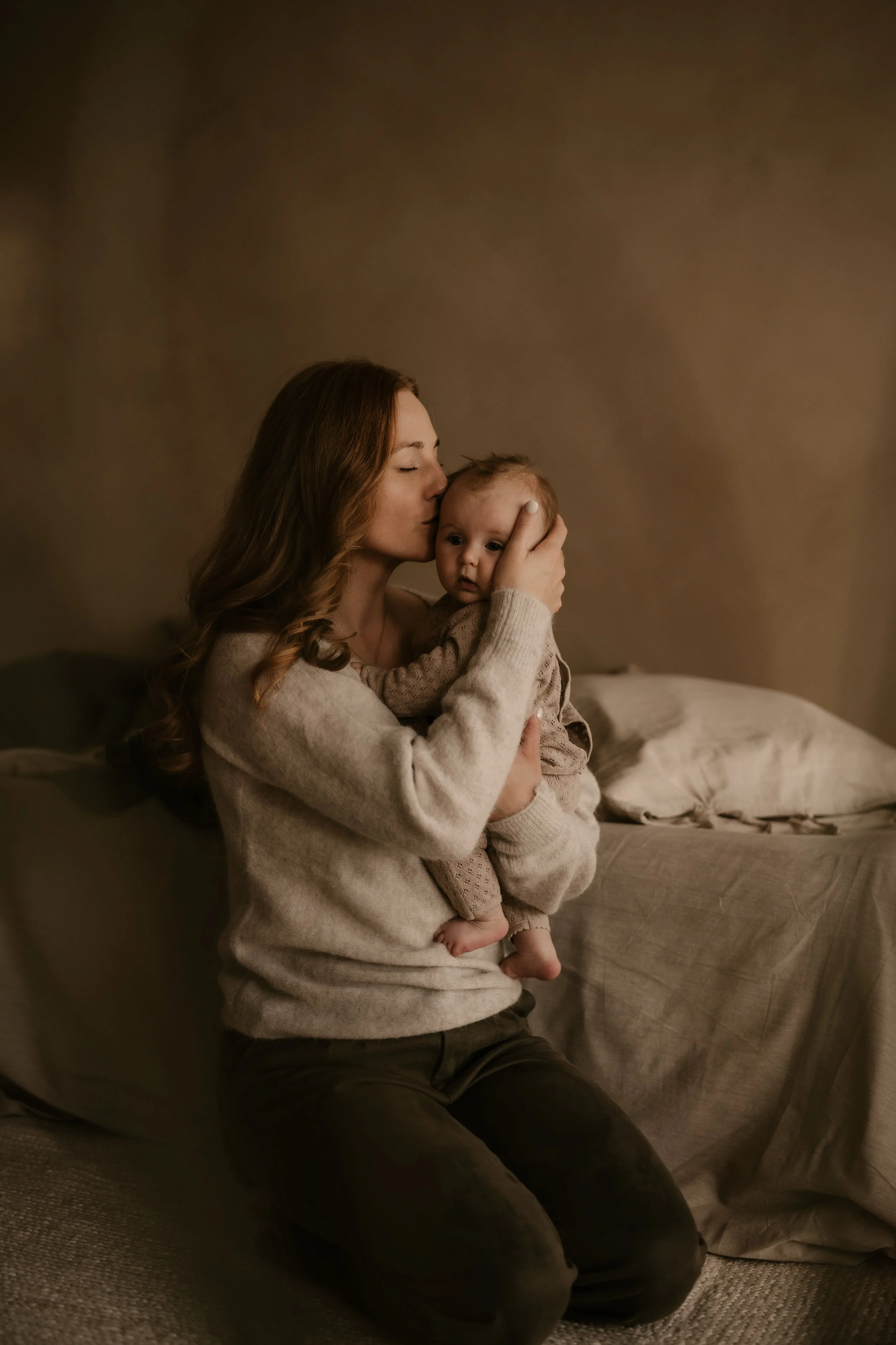 Eine Frau mit langen braunen Haaren hält ein Baby und küsst es auf die Stirn, im Hintergrund ein Bett mit beige Bettwäsche.