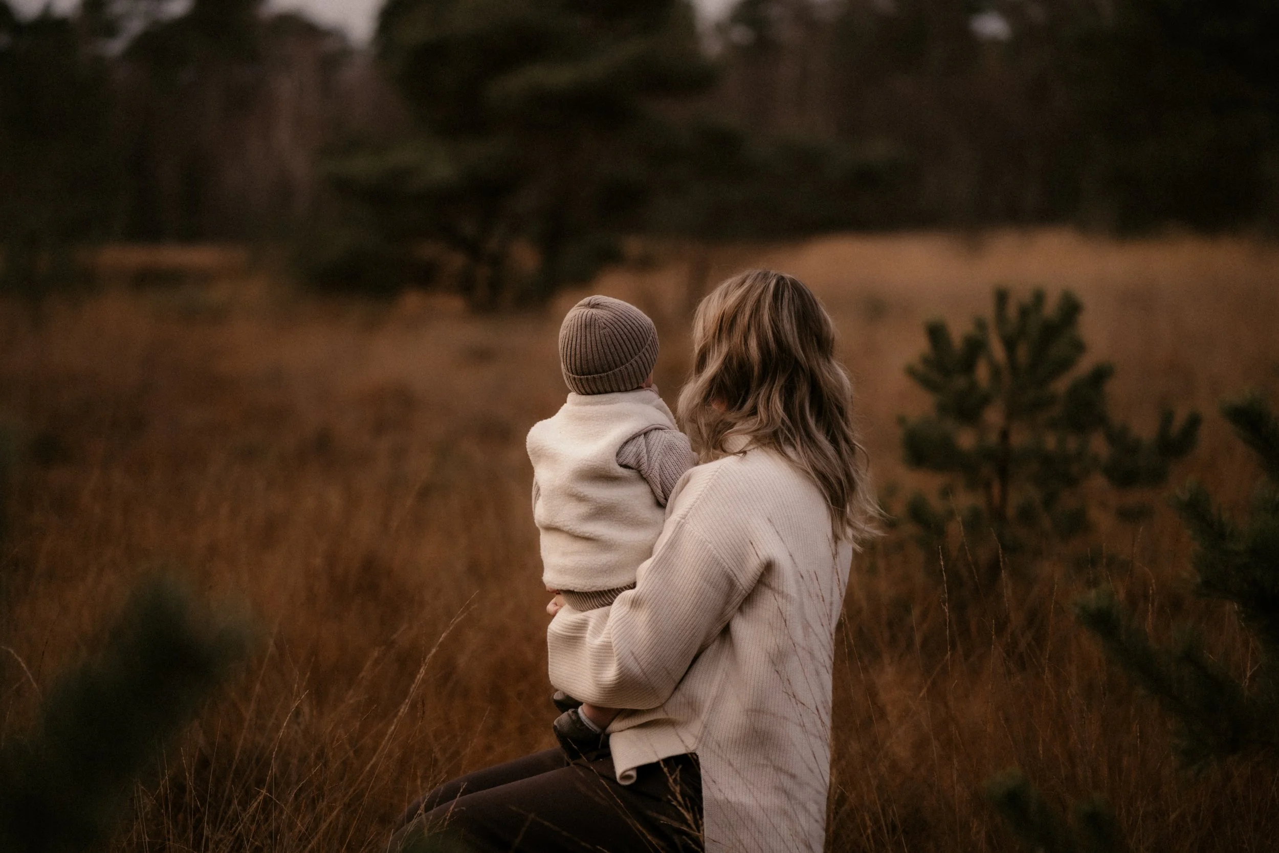 Eine Frau sitzt im Freien mit einem kleinen Kind auf ihrem Schoß, umgeben von wildem Gras und kleinen Tannenbäumen, bei Sonnenuntergang.