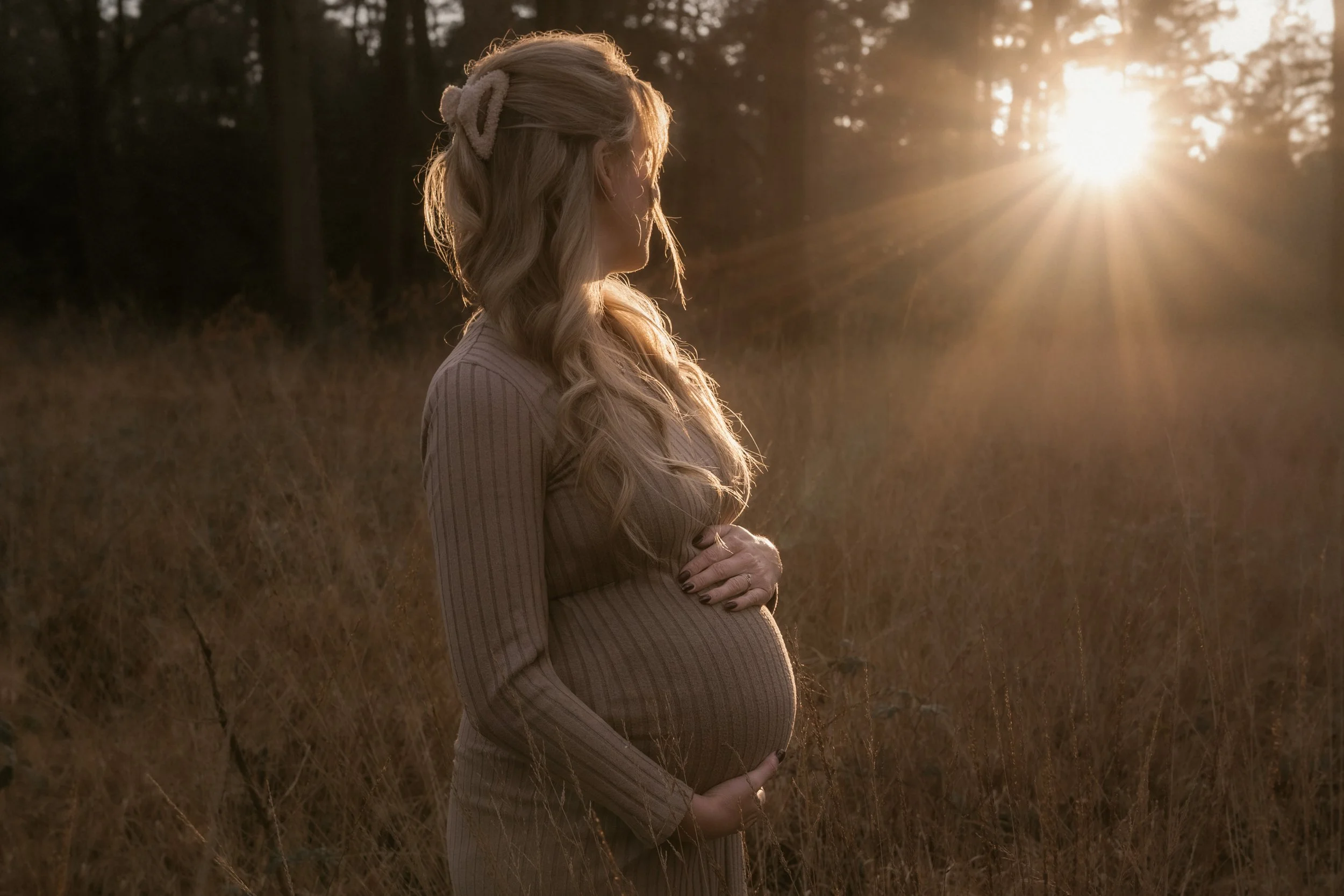 Eine schwangere Frau mit langem, gewelltem Haar steht in einem Feld bei Sonnenuntergang, die Sonne scheint durch die Bäume im Hintergrund.