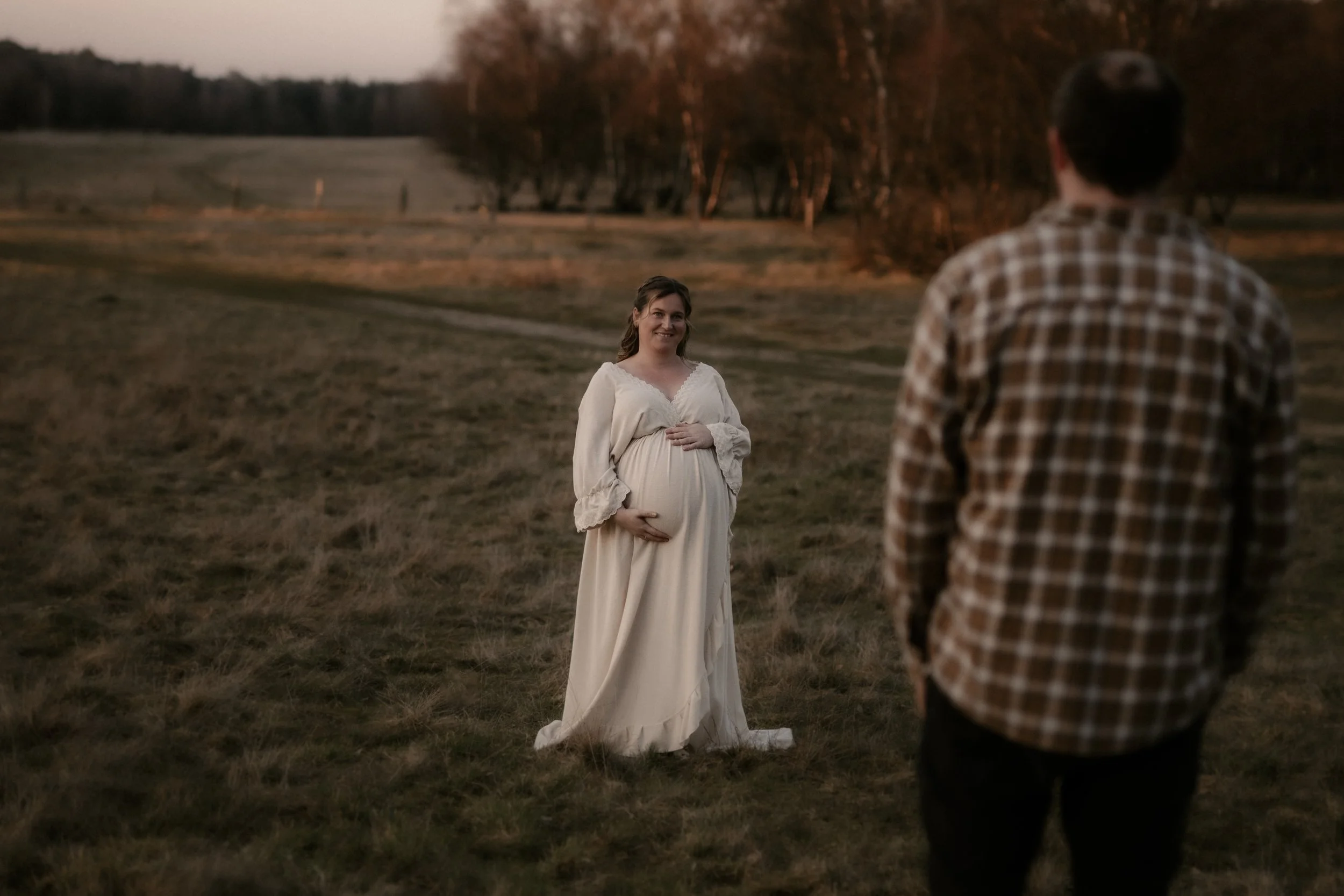 Ein schwangere Frau in einem weißen Kleid steht im Freien während des Sonnenuntergangs, während ein Mann in kariertem Hemd sie ansieht.