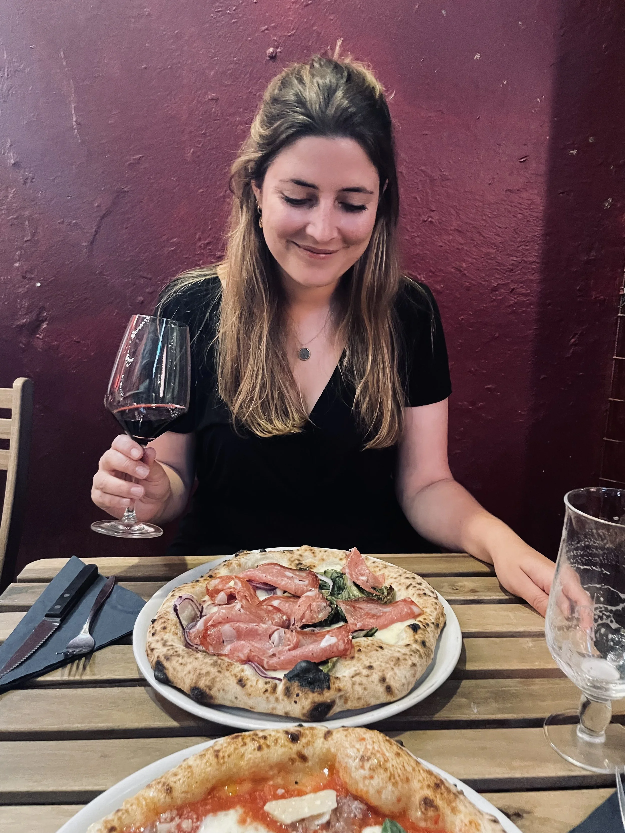 Eine Frau sitzt an einem Restauranttisch, hält ein Glas Rotwein und blickt auf eine Pizza mit Schinken und Zwiebeln.