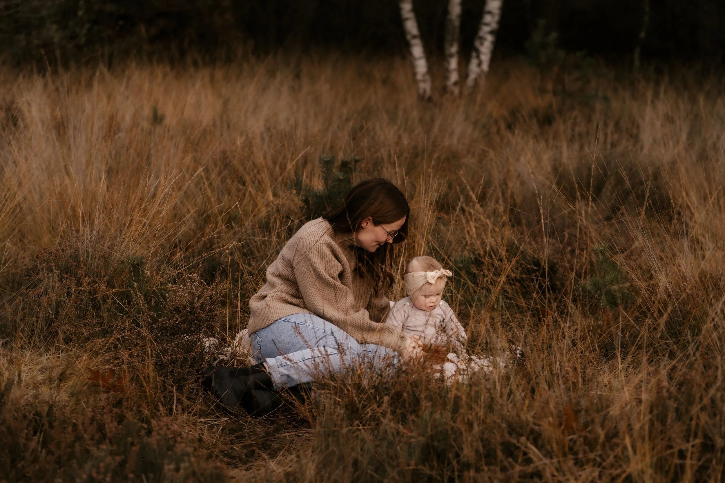 Eine Frau und ein Mädchen sitzen im hohen Gras in einem Waldstück, das blond und braun gefärbt ist. Die Frau trägt eine braune Strickjacke und Jeans, während das Mädchen ein Lätzchen und eine Schleife im Haar trägt.