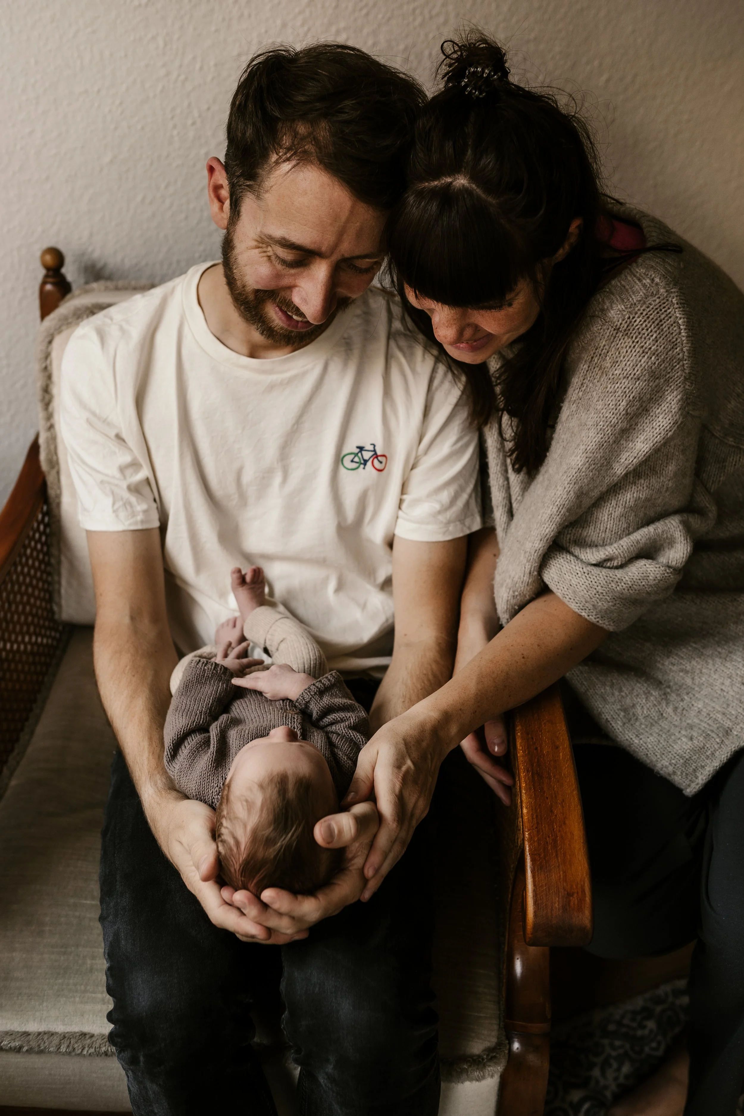 Ein Paar hält ein neugeborenes Baby auf dem Schoß und schaut liebevoll auf das Kind, das in ihren Armen liegt.