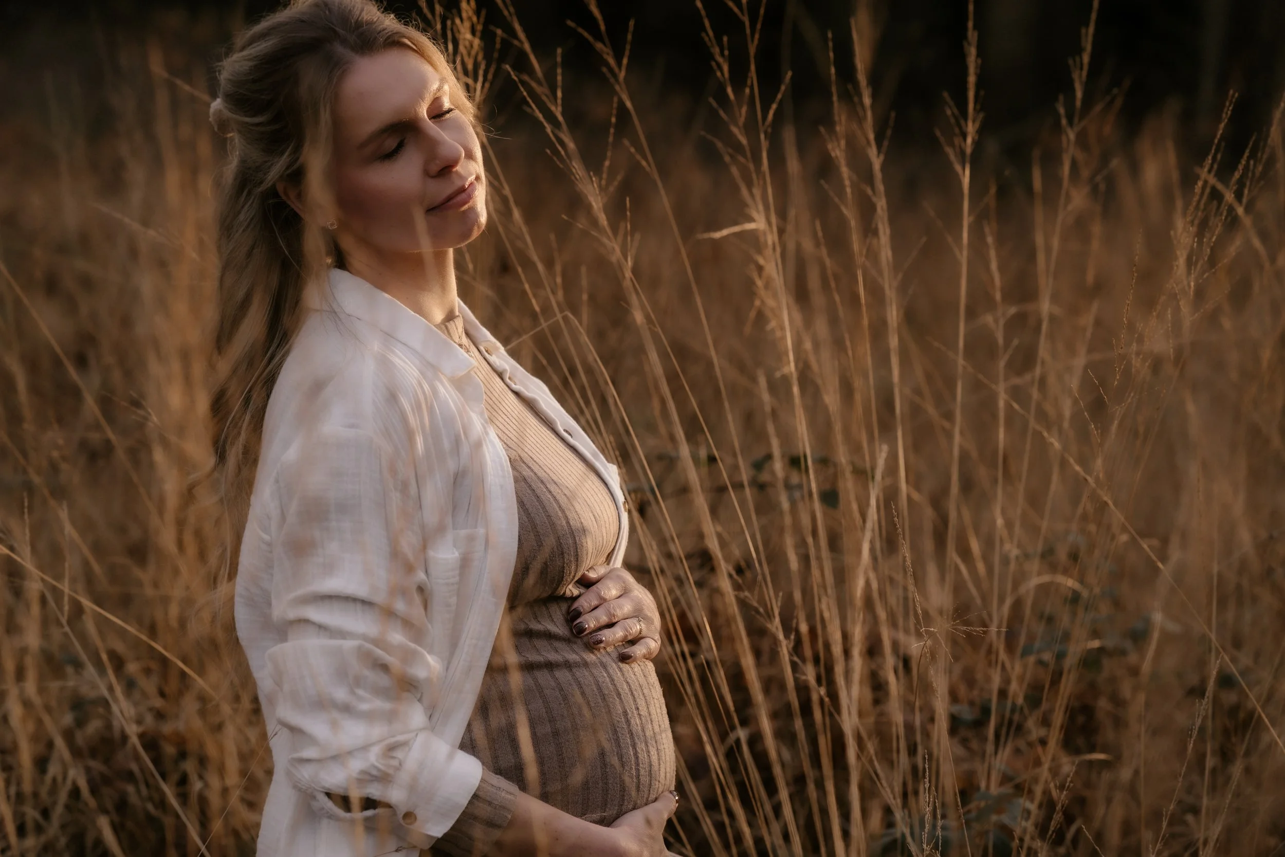 Eine schwangere Frau steht in einem Feld mit getrocknetem Gras, hat die Augen geschlossen und wirkt zufrieden, während sie ihre Schwangerschaft barem zeigt.