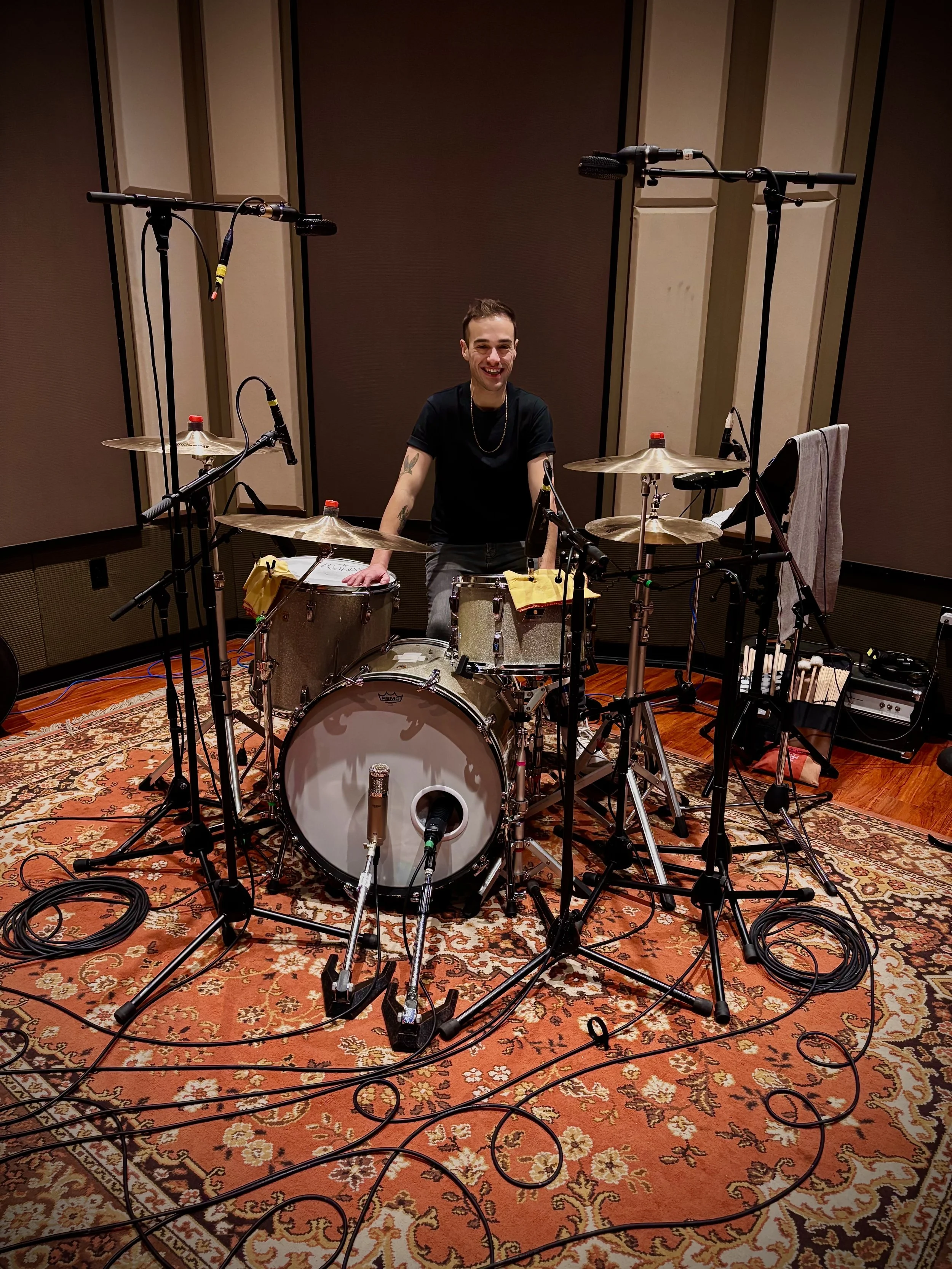 A man is sitting at a drum set in a recording studio, smiling. The drum set is surrounded by multiple microphones, and there are cables on the floor. The studio has a patterned rug and acoustic panels on the walls.