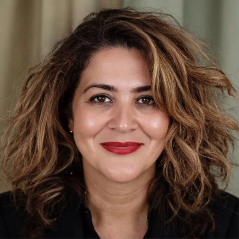 A woman with curly, shoulder-length brown hair with blonde highlights, smiling with red lipstick, wearing a black top.