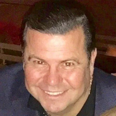 Close-up of a smiling man with short, dark hair, wearing a dark blazer and a black shirt, indoors.
