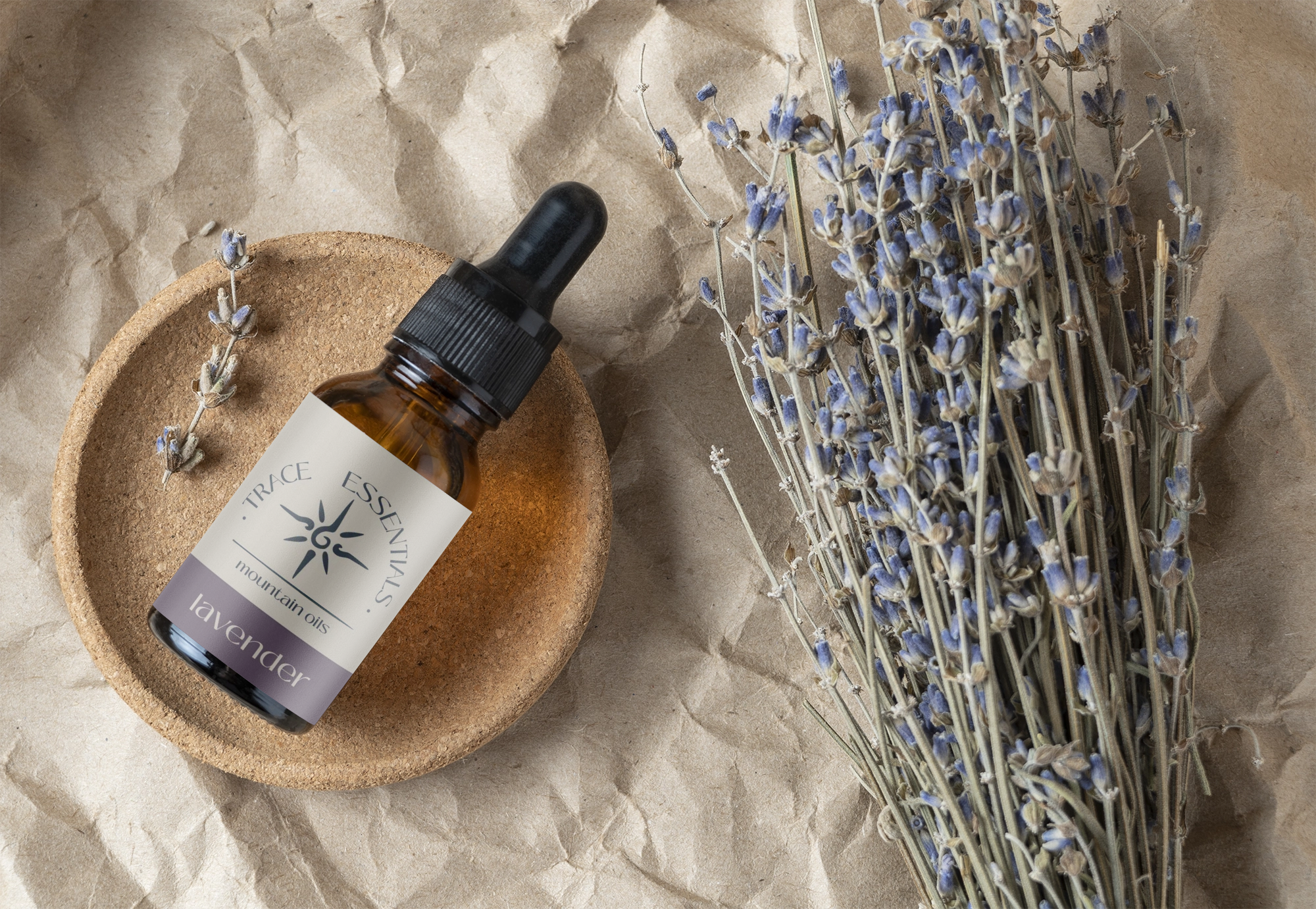 A small amber glass bottle of lavender essential oil with a black dropper cap placed in a round cork dish, alongside a bundle of dried lavender flowers on crinkled brown paper.