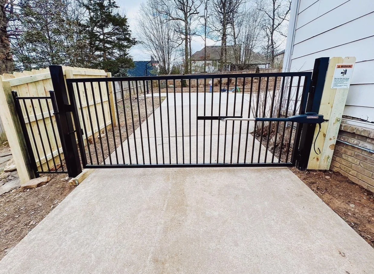 Automated black steel driveway gate with a gate opener arm and a matching walk gate beside a wood privacy fence.