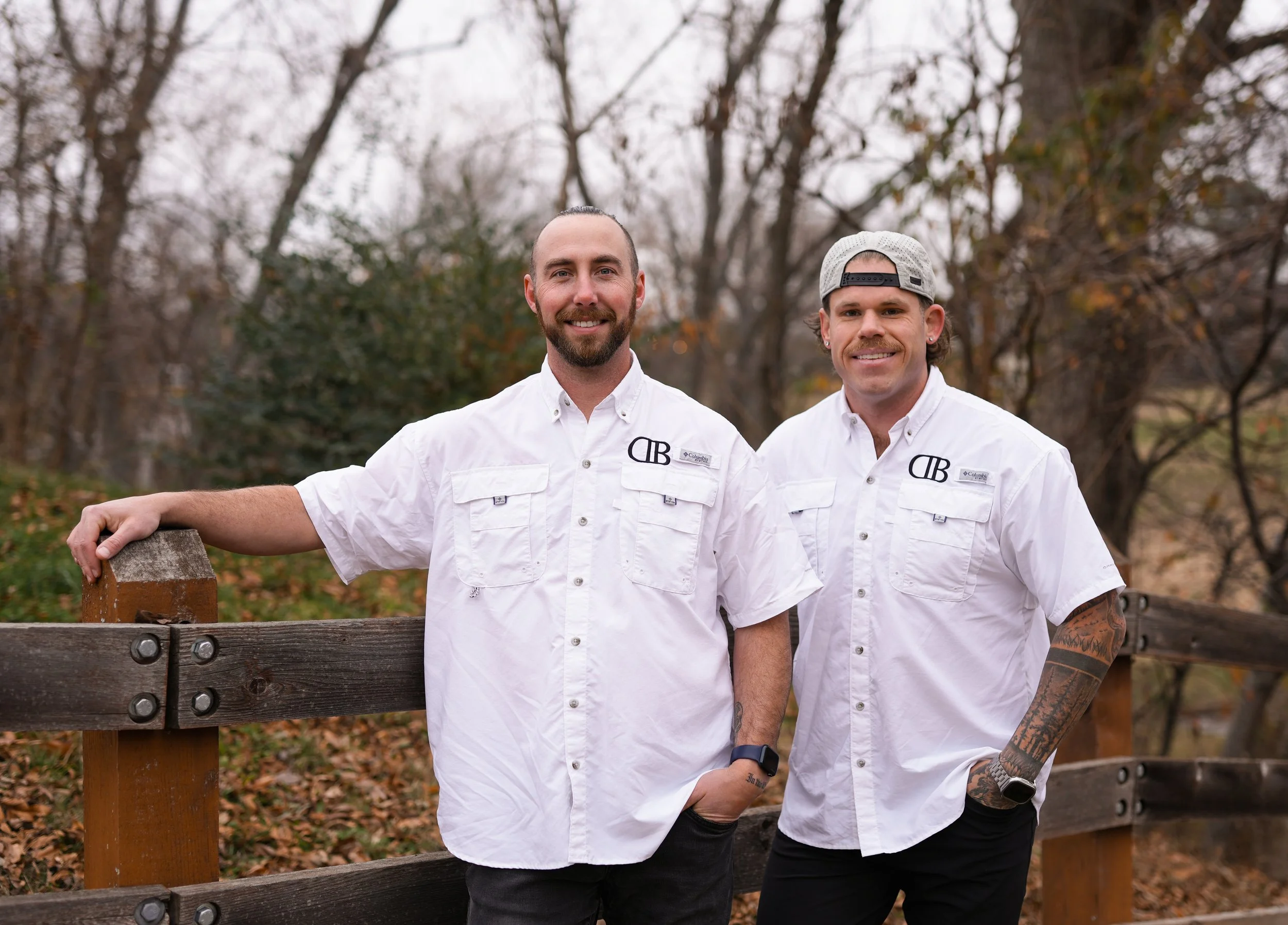 Two men with white shirts standing outdoors on a wooden bridge, with trees in the background and fall leaves on the ground.