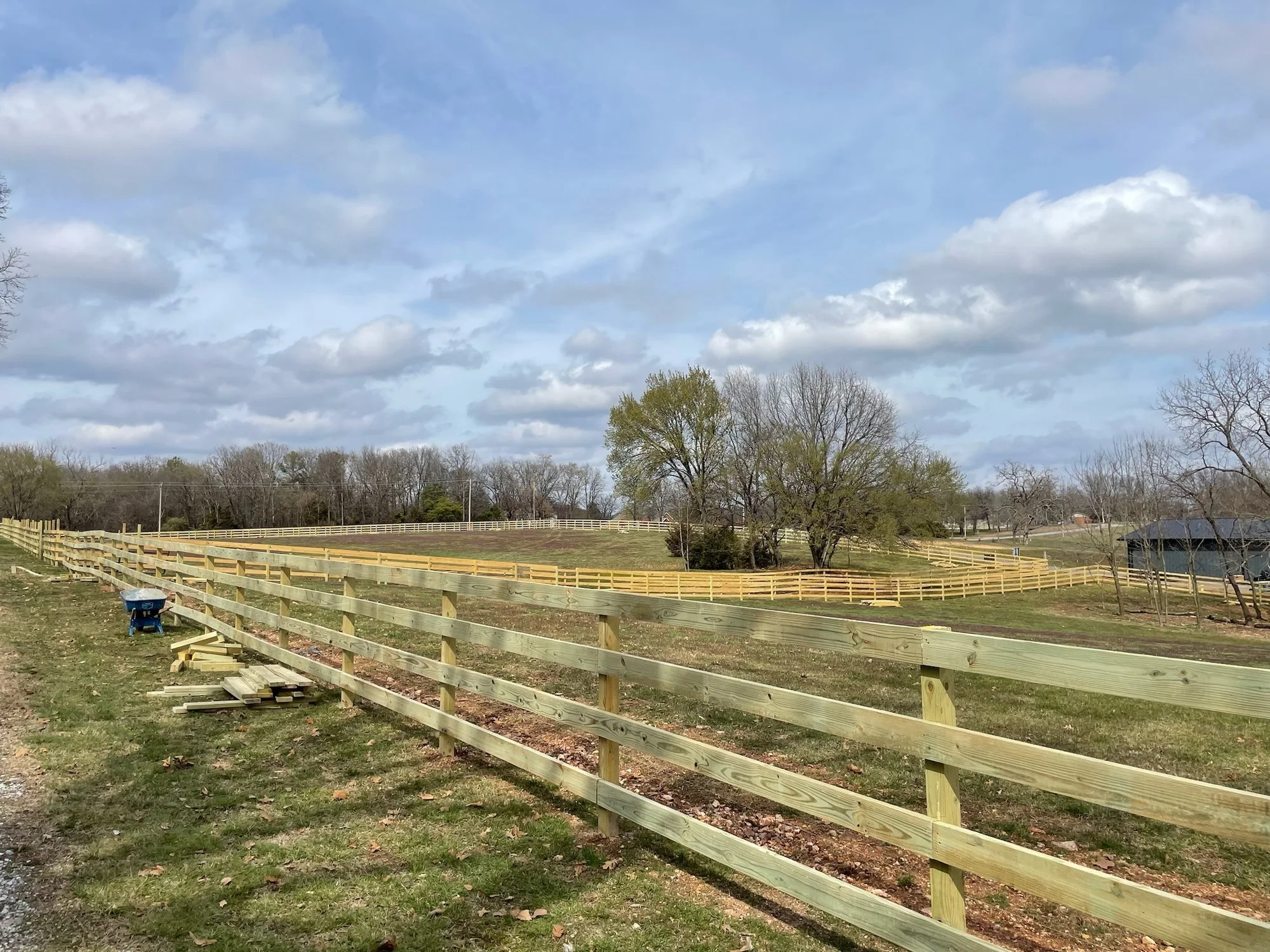 New four-board wood pasture fence running across open acreage, with a stack of lumber beside the fence line.