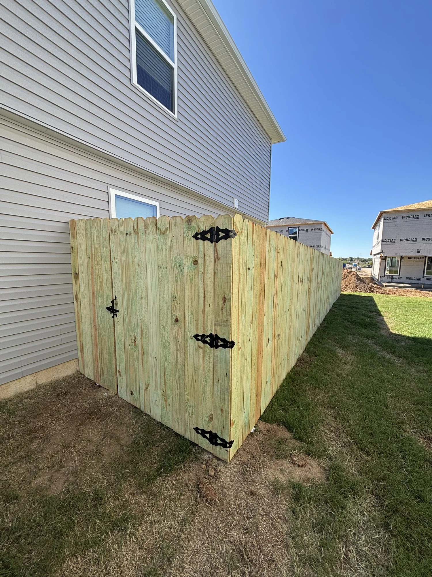 New pine privacy fence and walk gate with black strap hinges closing off a side yard between two homes.