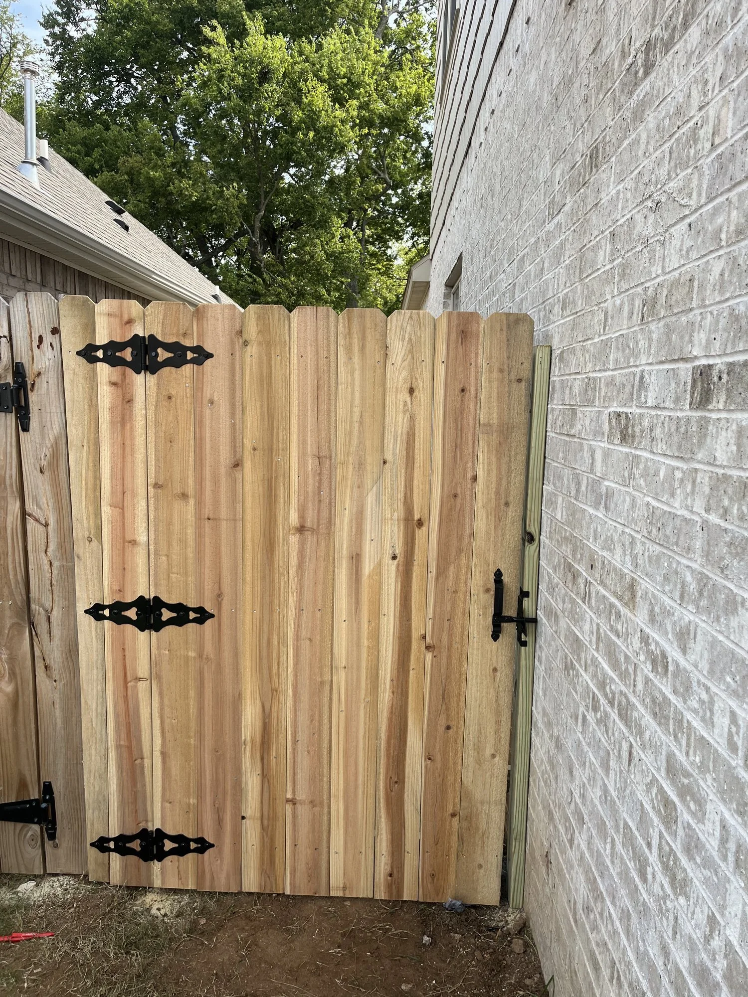 Natural wood privacy walk gate with black strap hinges and a black latch, next to a light brick wall.