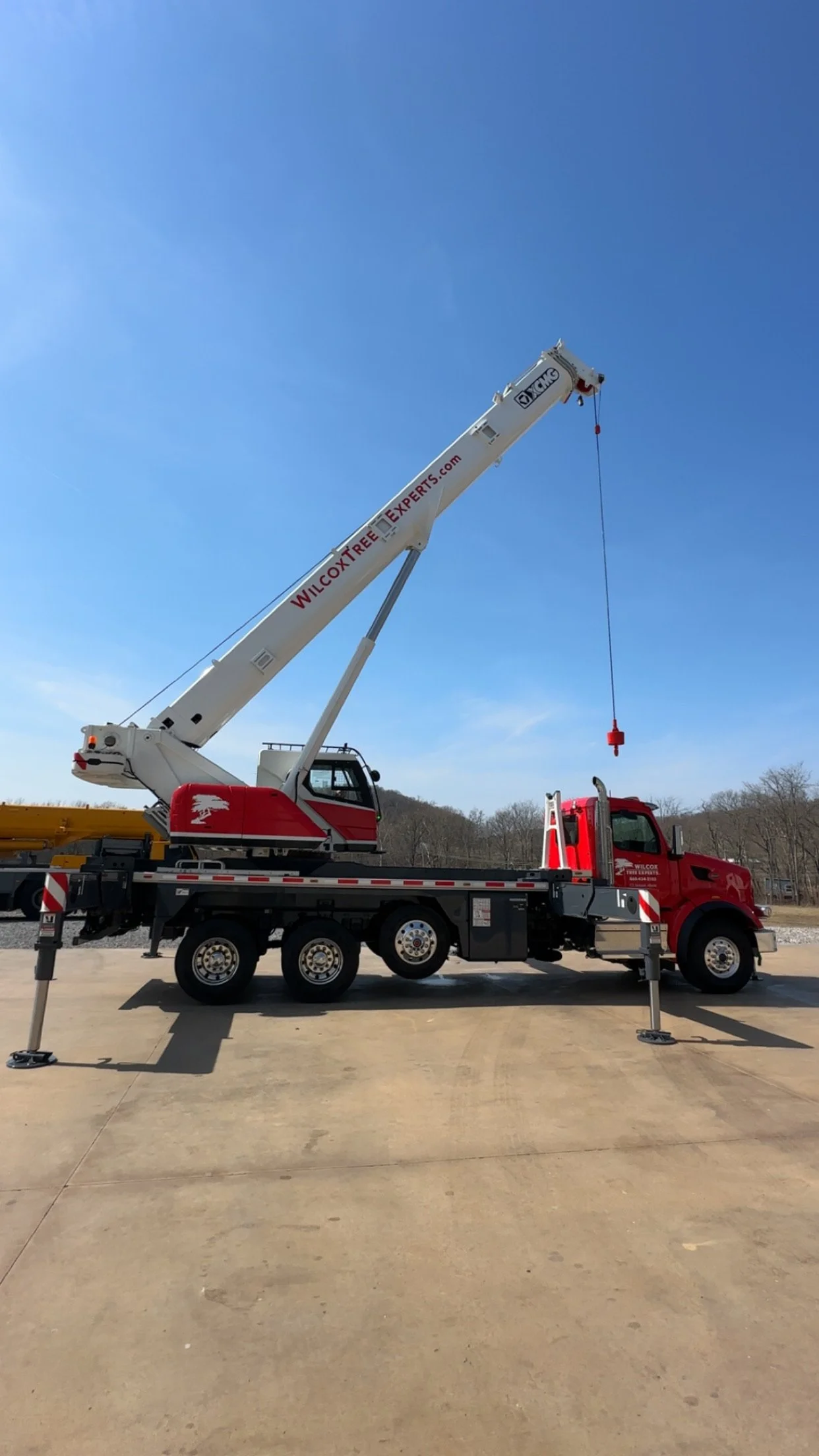 Red and white crane truck with a long boom and a hook hanging from it, parked on a concrete surface outdoors under a clear blue sky.
