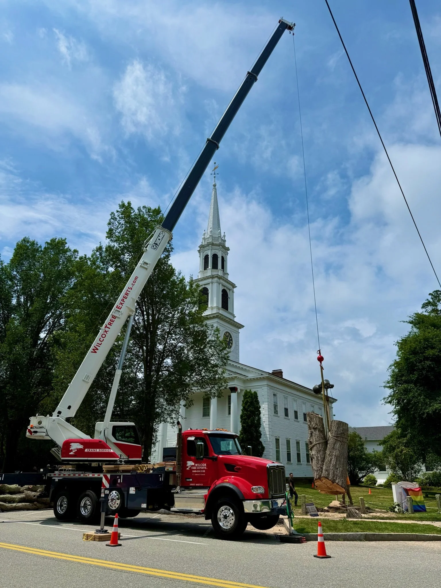 Tree removal in progress with a crane truck in front of a white church with a tall steeple, clear blue sky, and traffic cones around the work area.