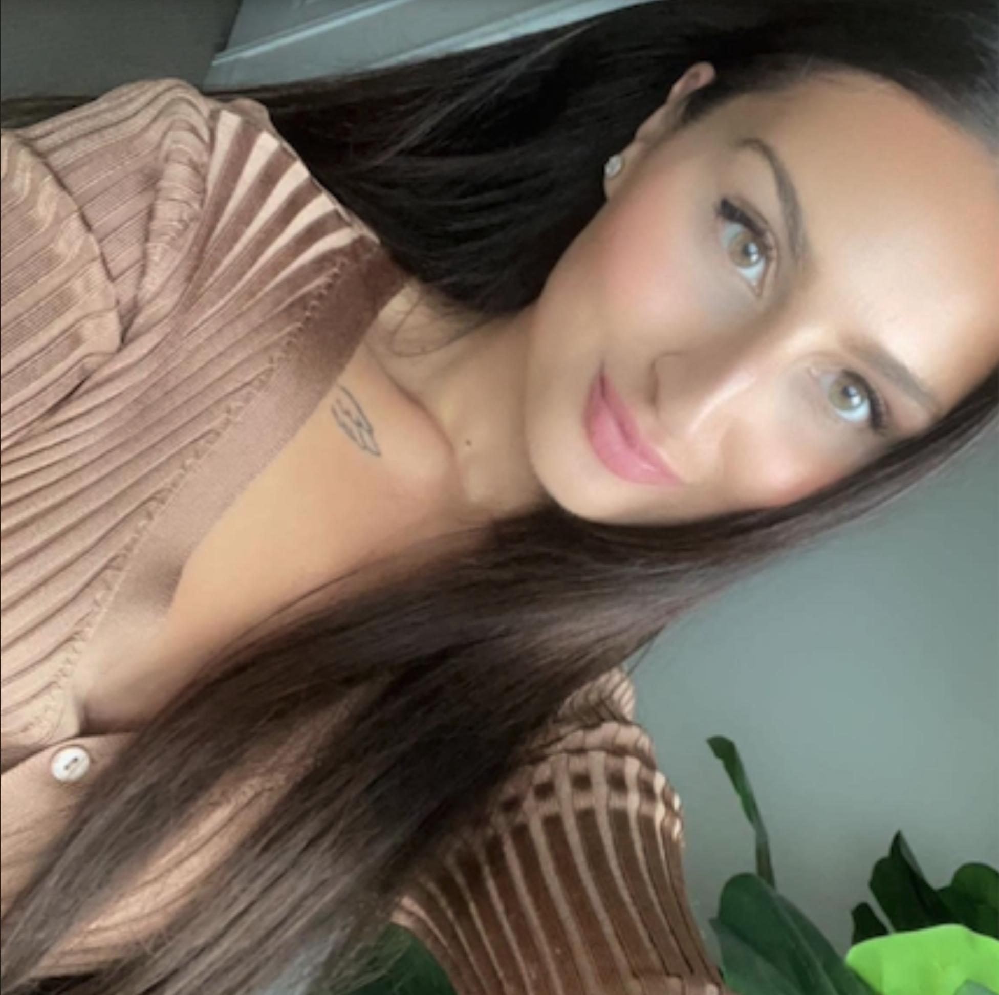 A woman with long dark hair, wearing a striped brown top, smiling softly with pink lipstick, in front of a plant with green leaves.