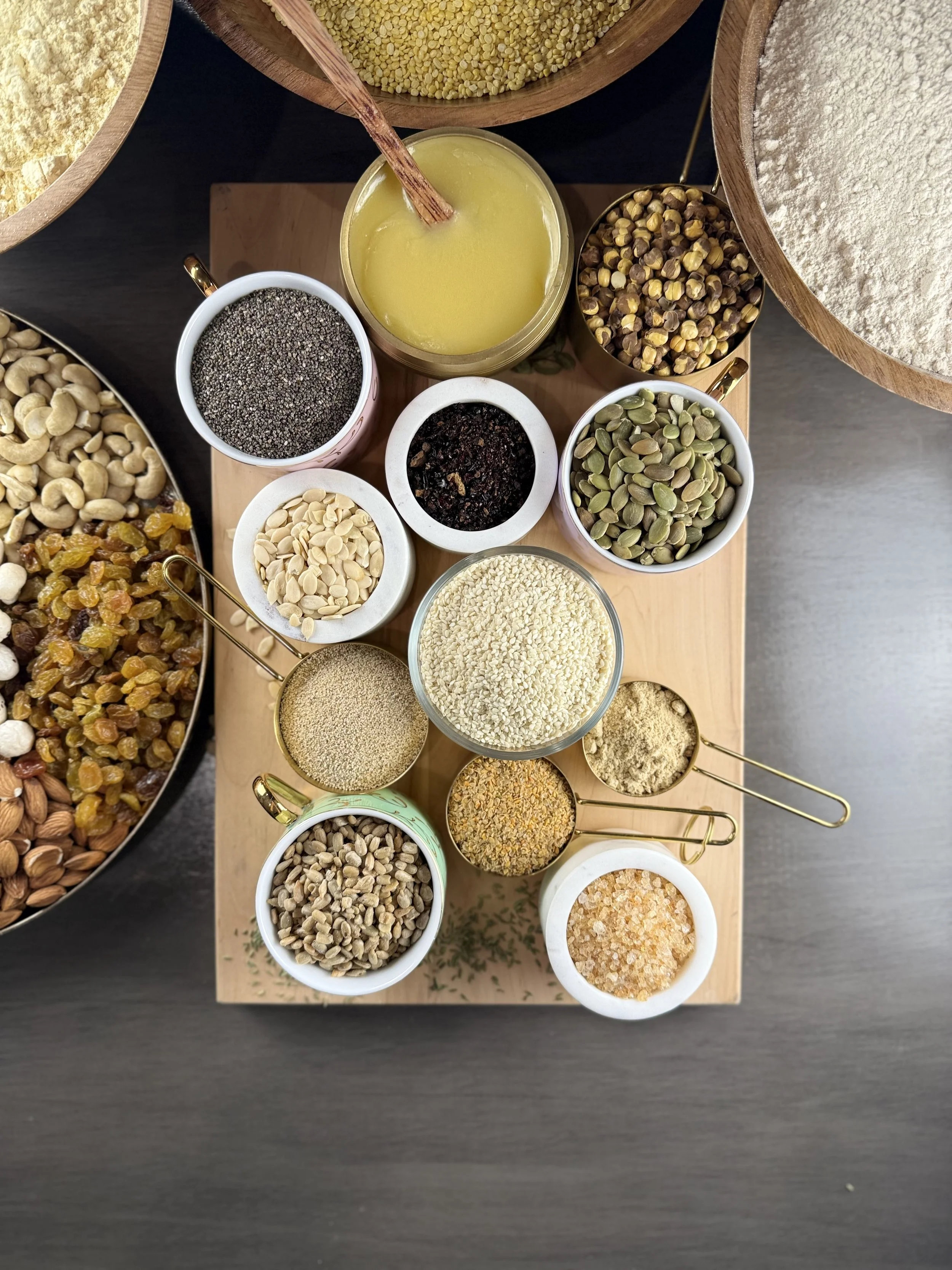 An assortment of bowls and jars filled with various seeds, nuts, and grains on a wooden board and black surface, including pumpkin seeds, sesame seeds, chia seeds, turmeric, and flaxseeds.