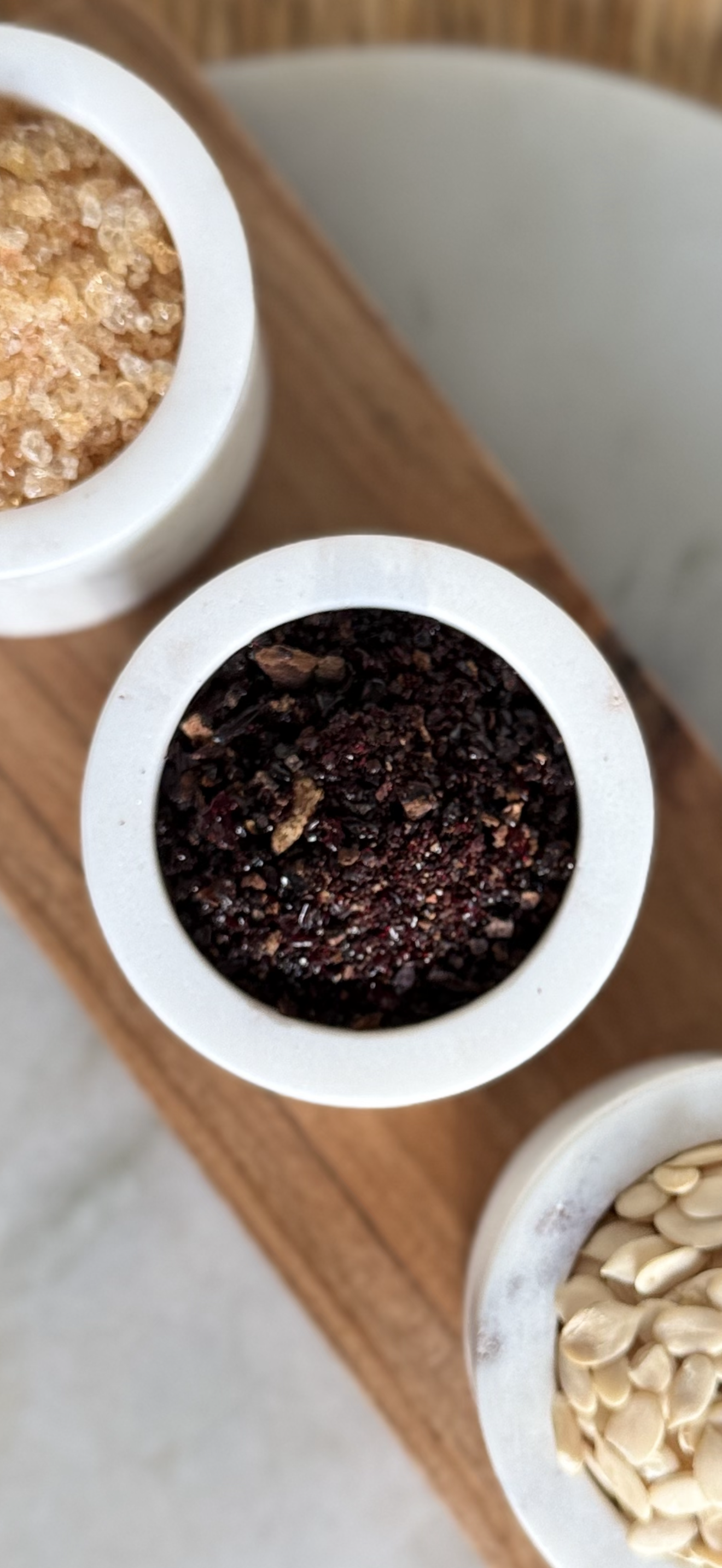 Close-up of three small white containers on a wooden surface, filled with pink salt, black pepper, and white sesame seeds.